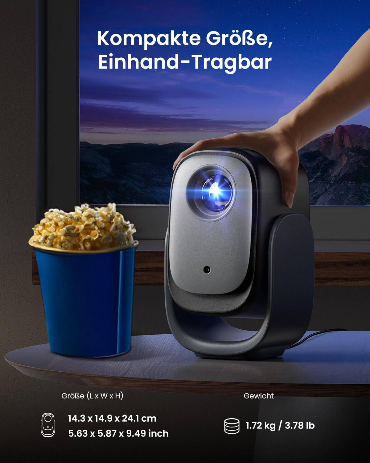 Una mano tiene un proiettore compatto su un tavolo accanto a un secchio di popcorn blu davanti a un paesaggio.