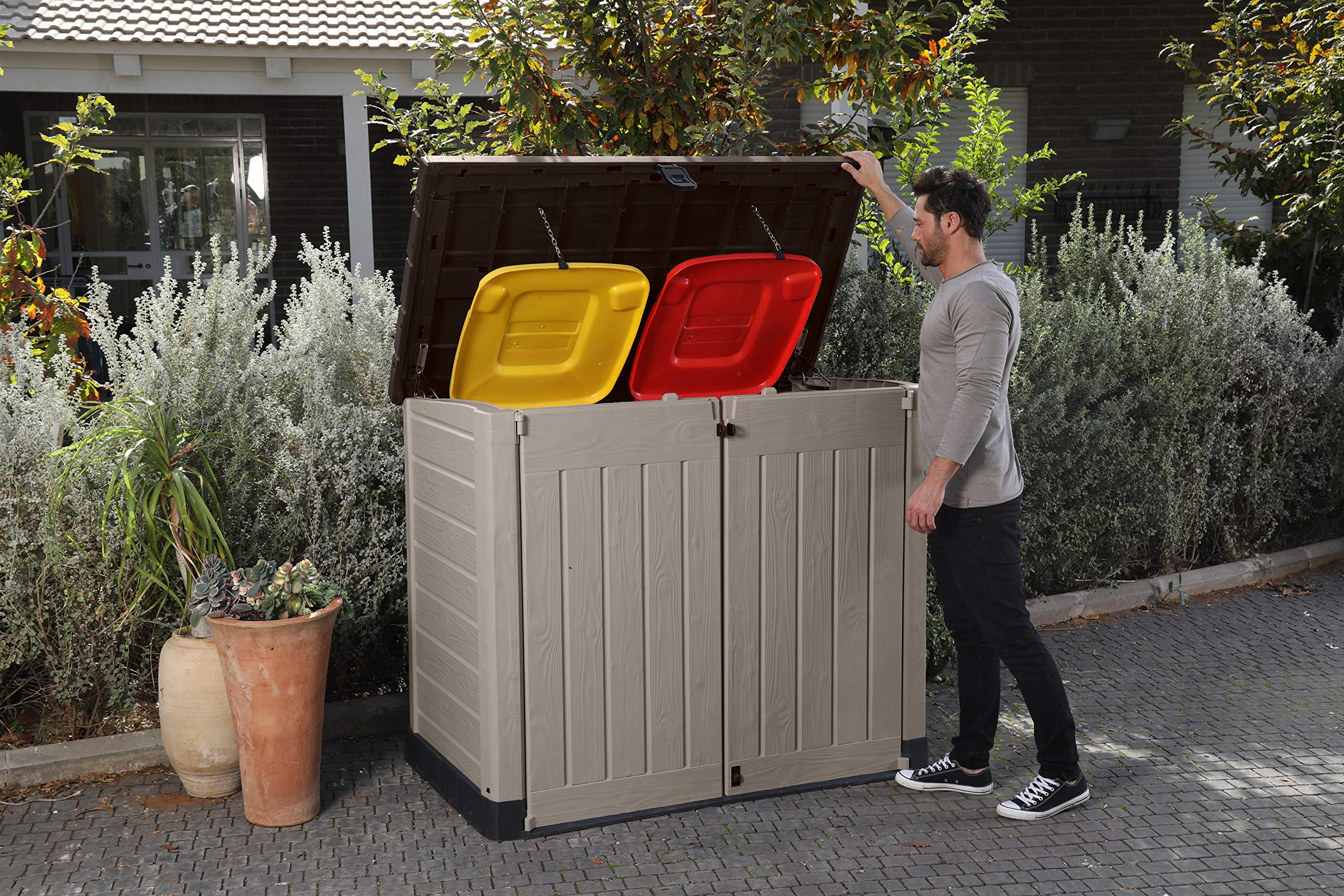 Ein Mann öffnet auf einer Terrasse einen Schuppen mit gelben und roten Mülleimern.