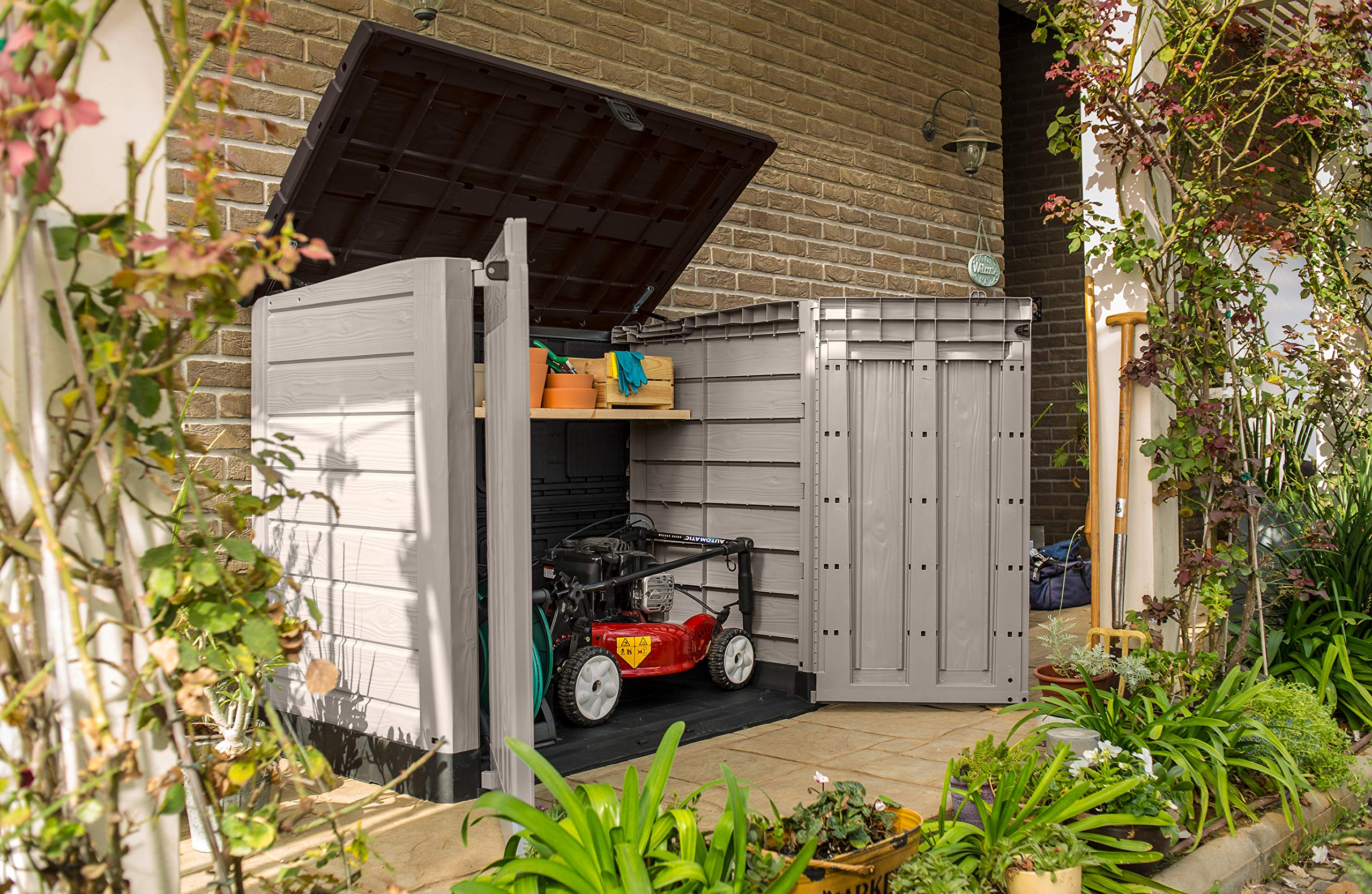 Ein grauer Kunststoff-Gartenschrank mit offenem Deckel und Türen steht auf einer Terrasse, Inhalt: Rasenmäher.