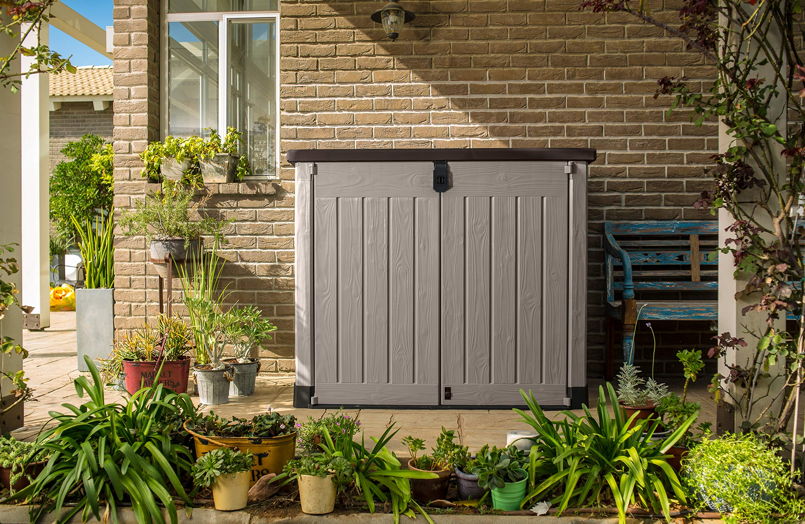 Eine graue Aufbewahrungsbox steht auf einer Terrasse vor einer Backsteinwand, umgeben von Topfpflanzen und einer blauen Bank.