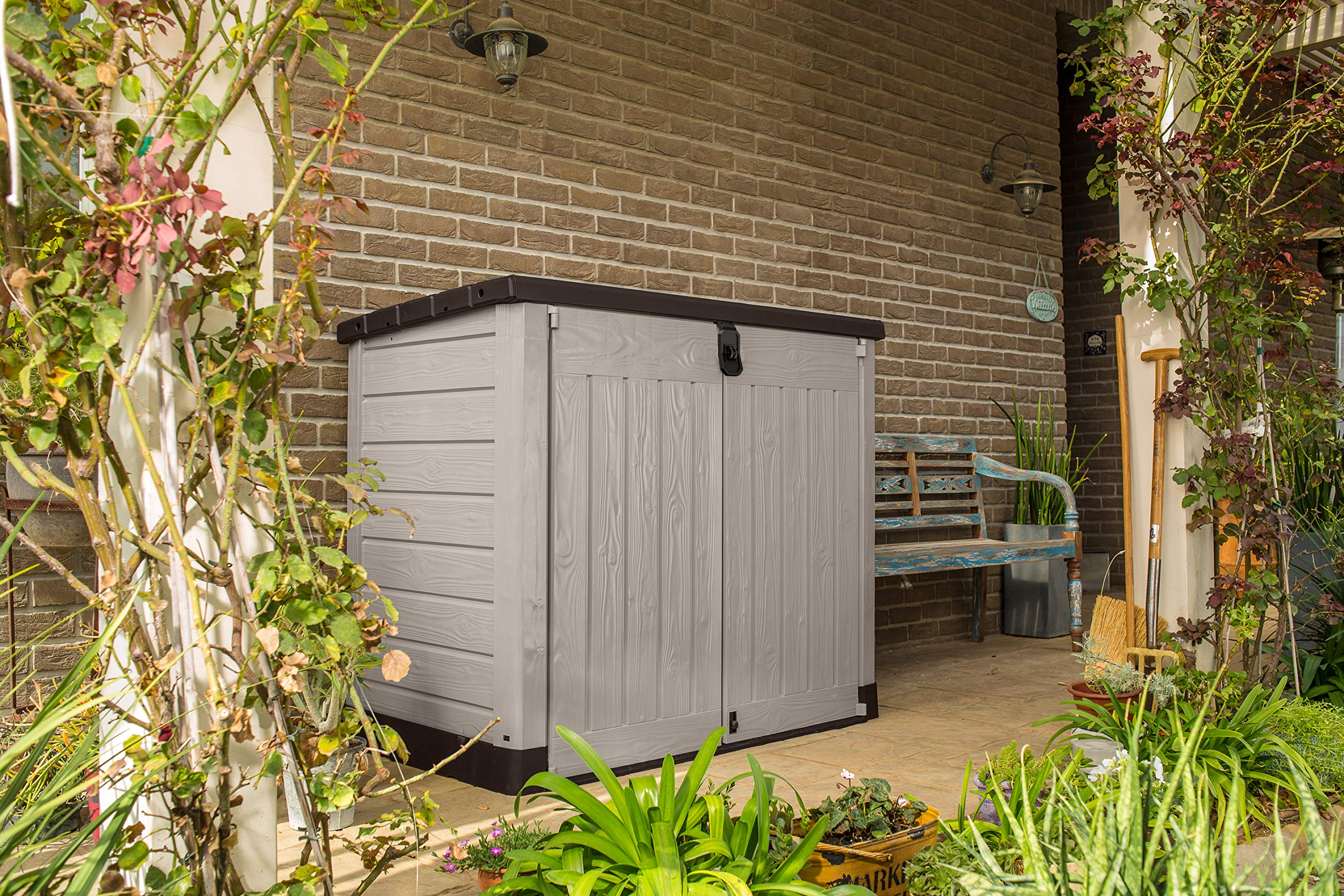 Eine graue Aufbewahrungsbox steht auf einer Terrasse vor einer Backsteinwand, umgeben von Pflanzen und einer Bank.