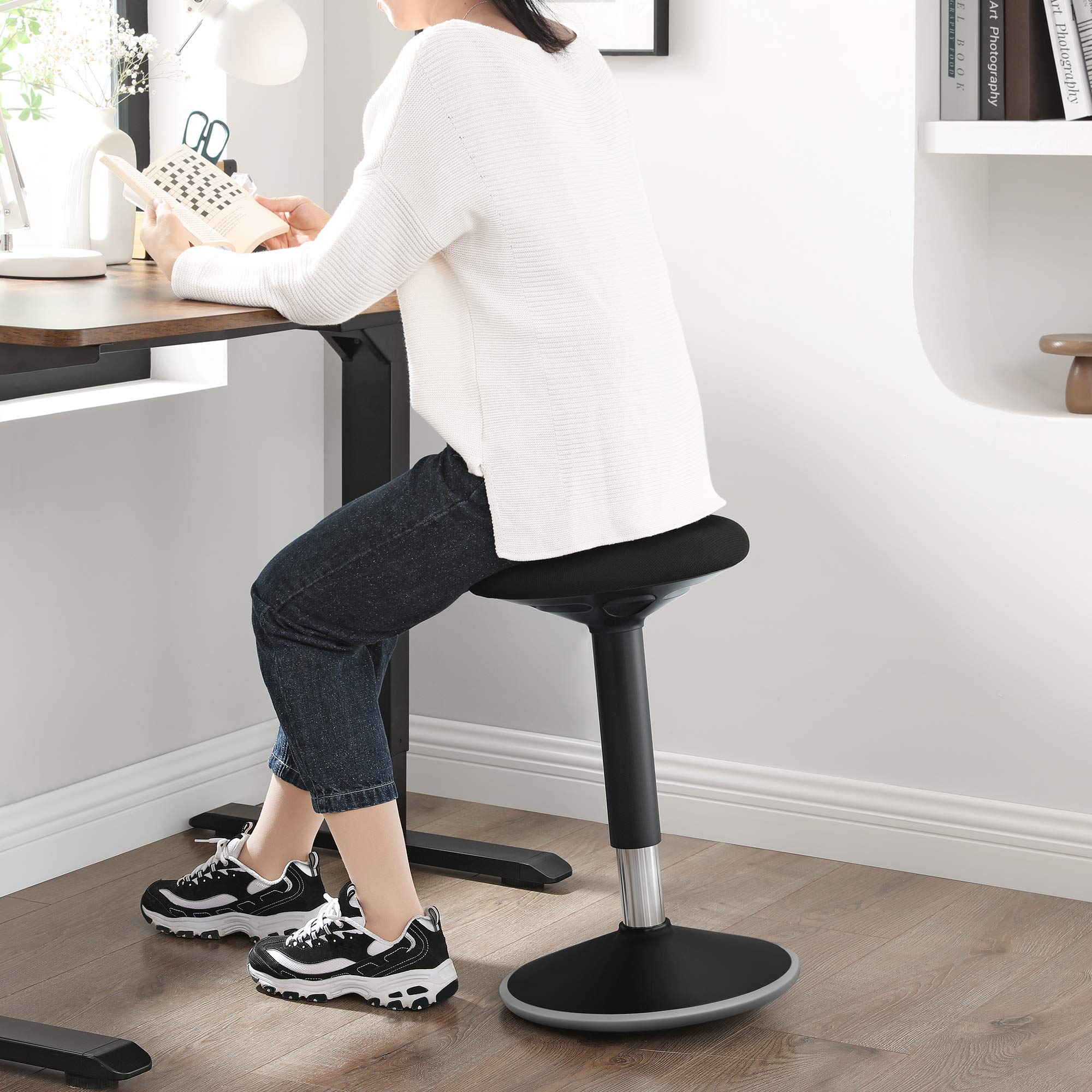 Une femme est assise sur un tabouret ergonomique noir à un bureau, lisant un livre.