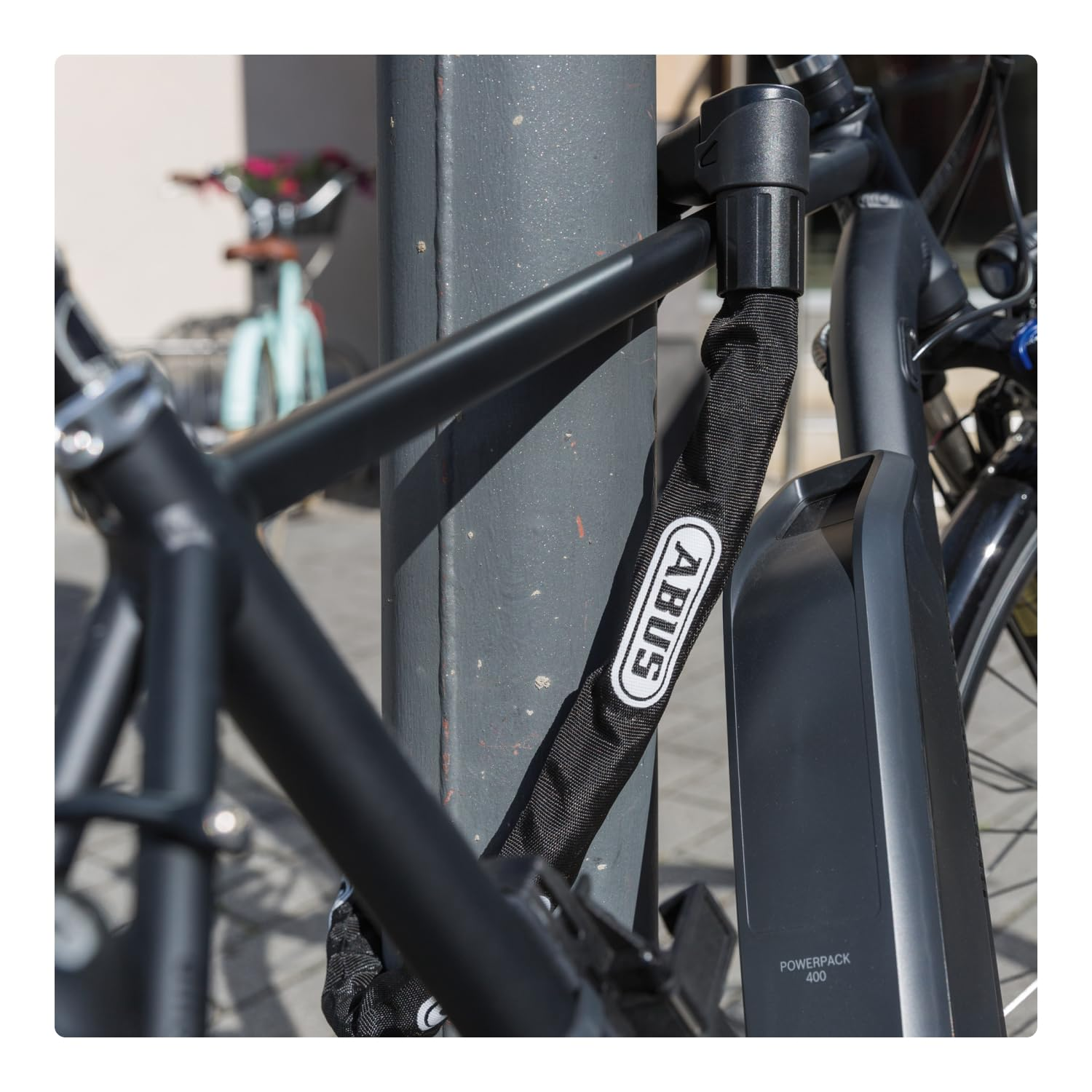 Un vélo  électrique noir est attaché à un poteau gris avec un antivol à chaîne Abus.