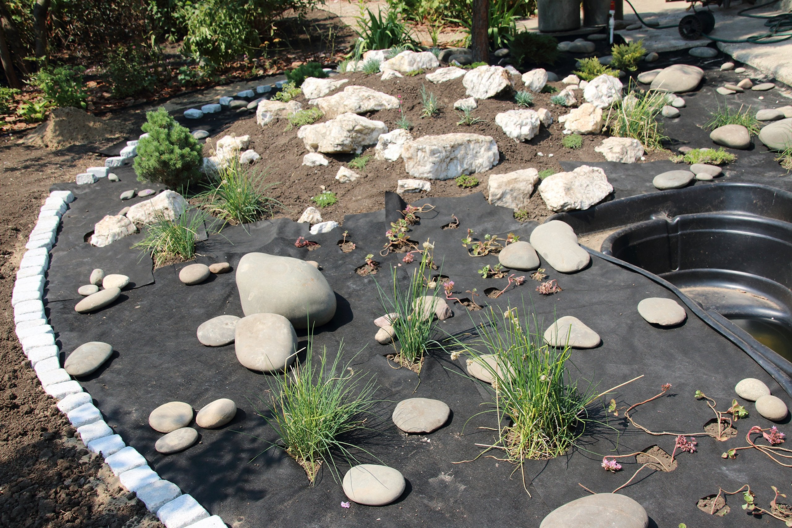 Un giardino in costruzione con telo nero antiradice, rocce decorative, piccole piante e un laghetto.