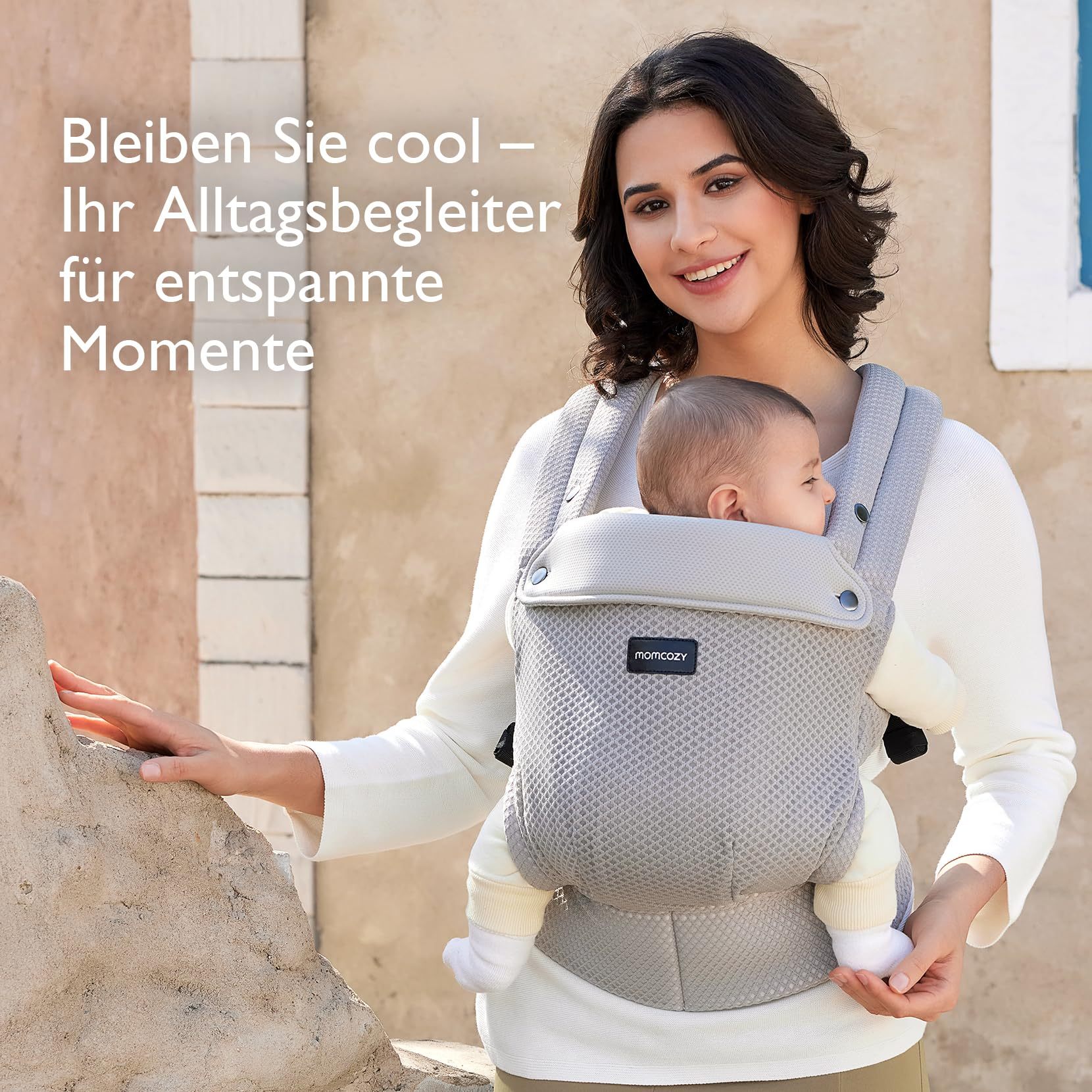 Una donna sorridente porta un bambino in un marsupio grigio contro una parete calda e strutturata.