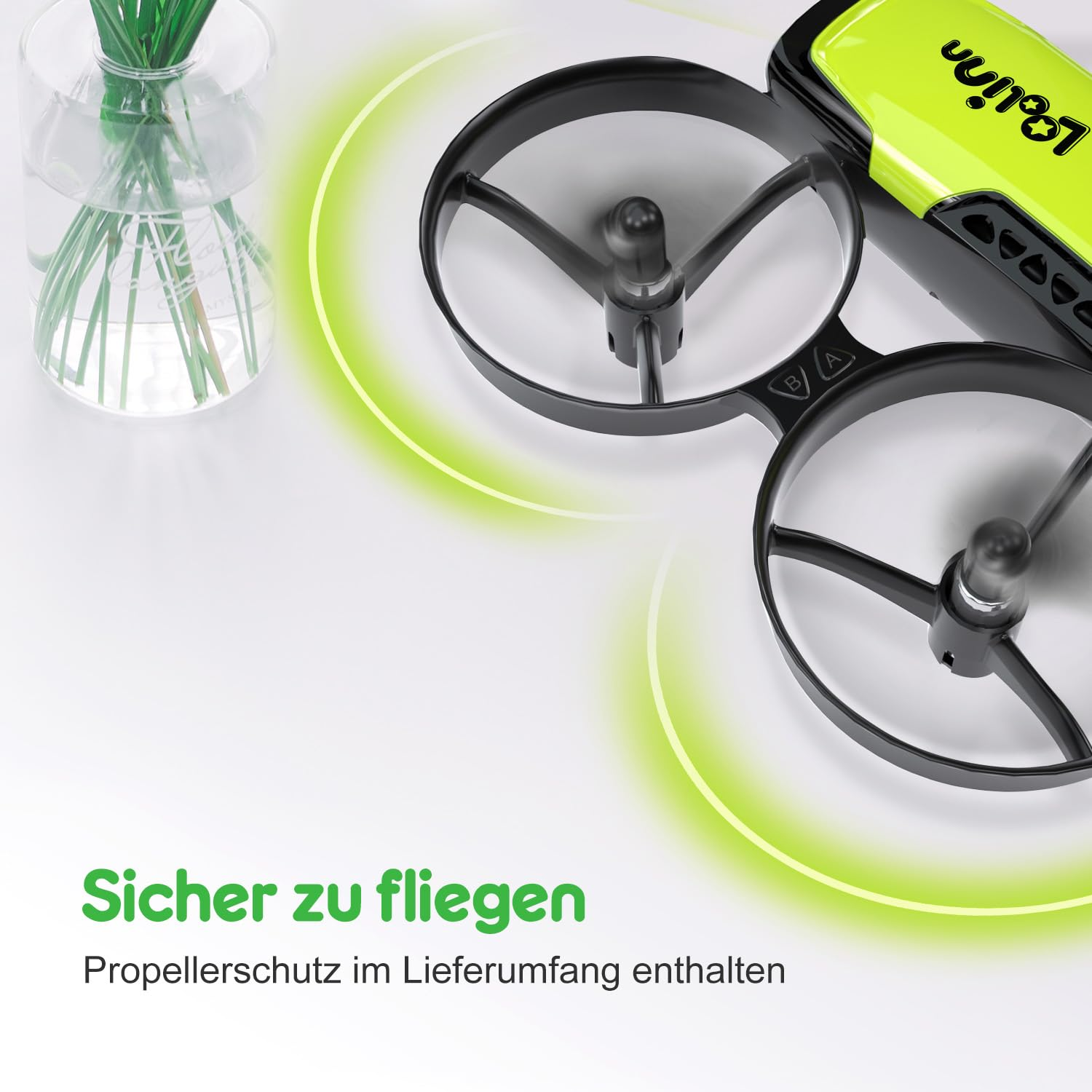 Primo piano di un drone verde e nero con protezioni per eliche su una superficie bianca, accanto a un vaso di vetro.