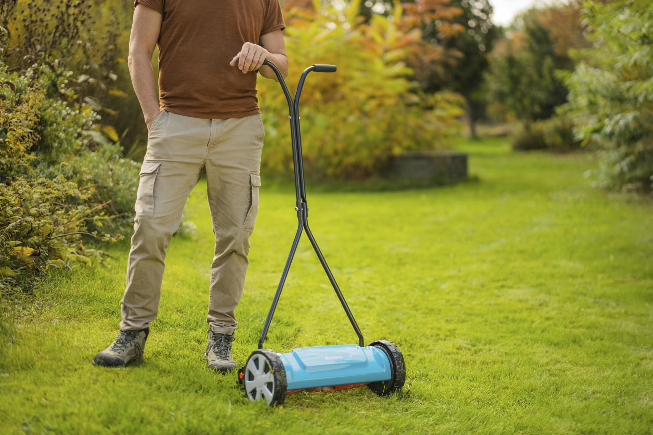 Un homme en t-shirt marron et pantalon kaki se tient dans un jardin à côté d'une tondeuse manuelle bleue.