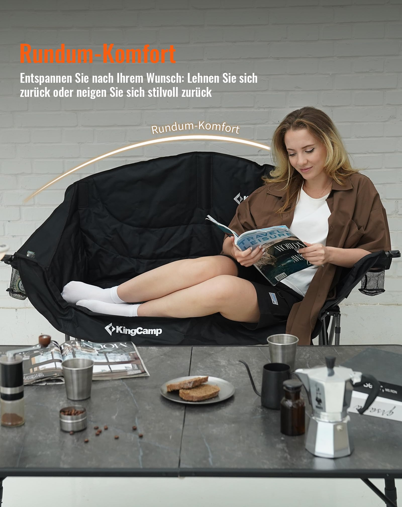 Eine Frau sitzt entspannt auf einem großen schwarzen KingCamp-Campingstuhl und liest eine Zeitschrift vor einer weißen Backsteinwand.
