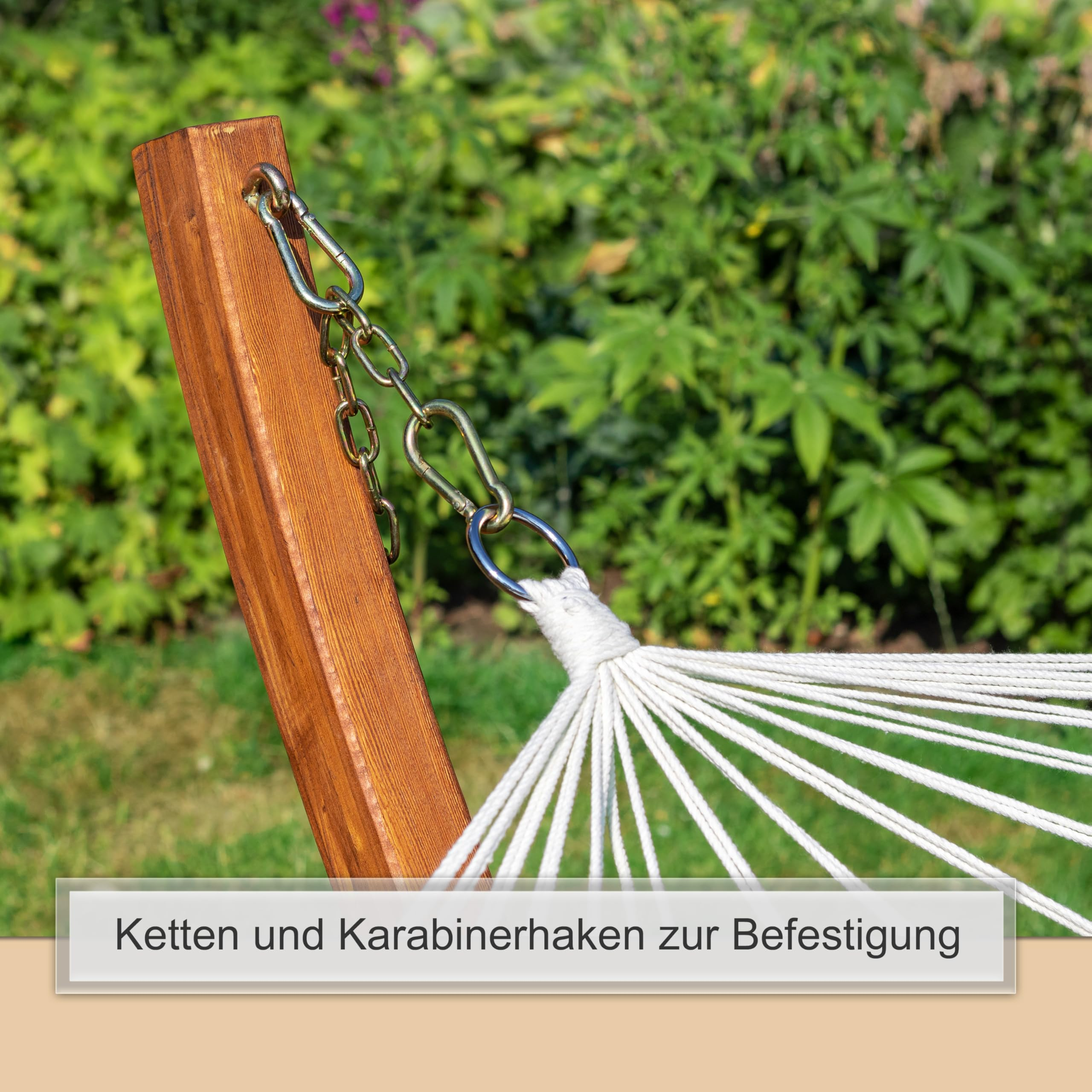 Weiße Seile einer Hängematte sind über Metallketten und Karabiner an einem Holzmasten im Garten befestigt.
