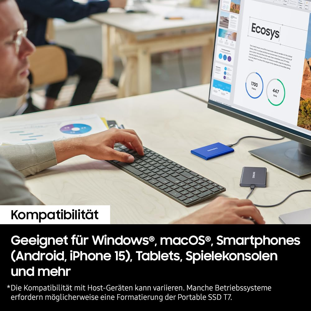 Una persona digita su una tastiera accanto a un monitor, con due SSD esterni e documenti su una scrivania.