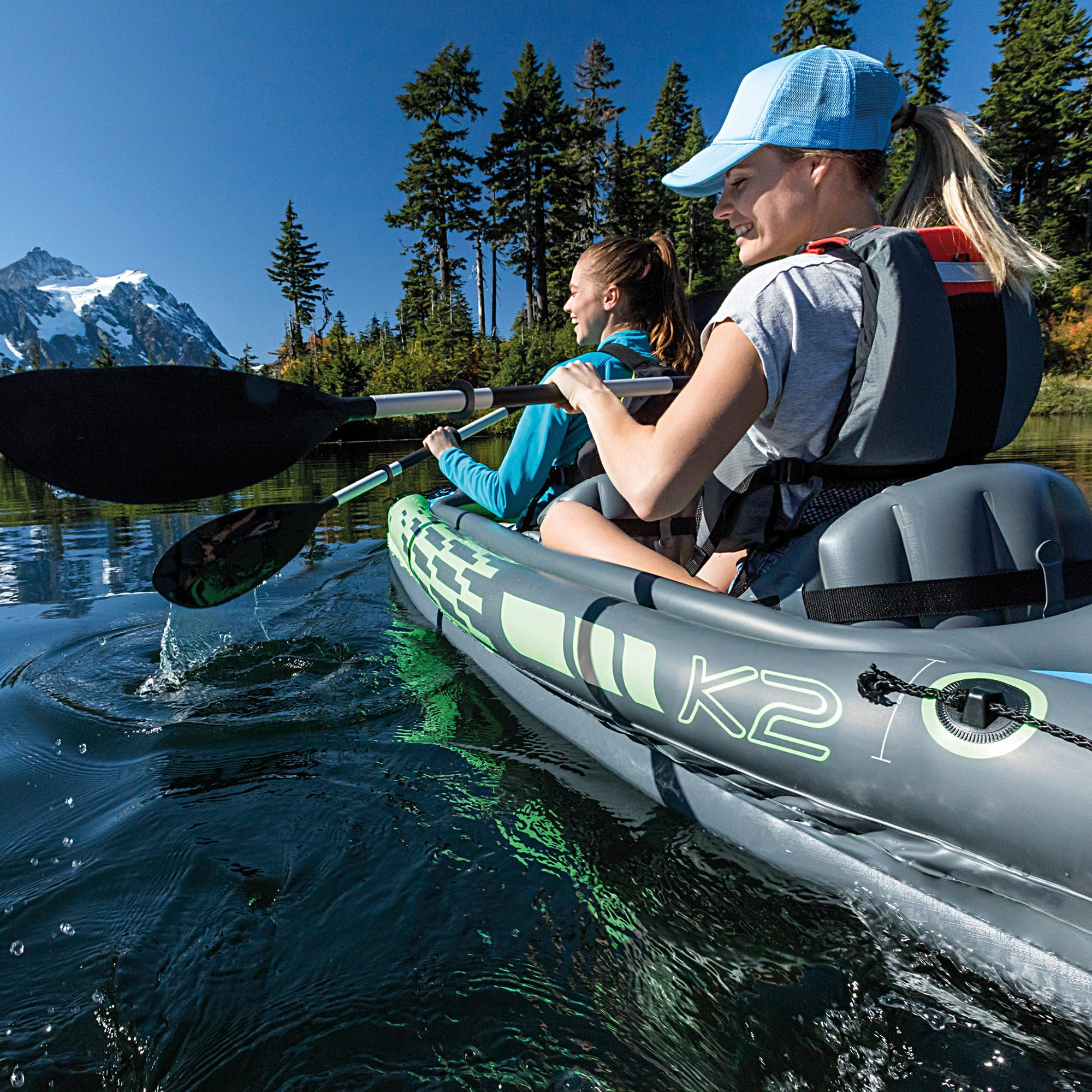 Zwei Frauen paddeln in einem grauen Inflatable-Kajak auf einem See vor einer Bergkulisse.