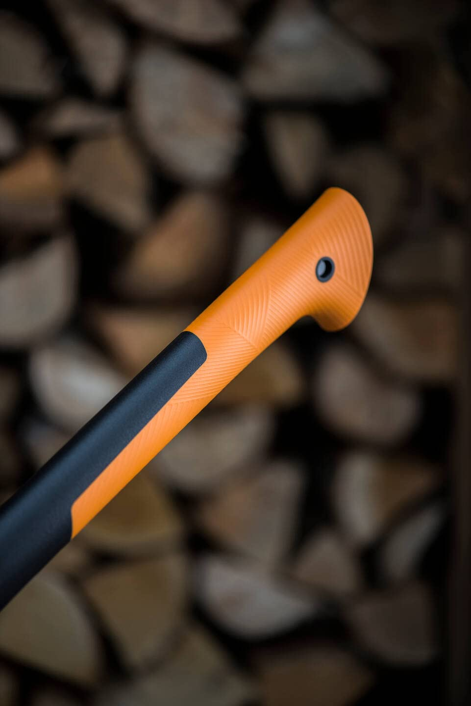 Nahaufnahme eines orangen und schwarzen Axtstiels vor einem verschwommenen Stapel Brennholz.