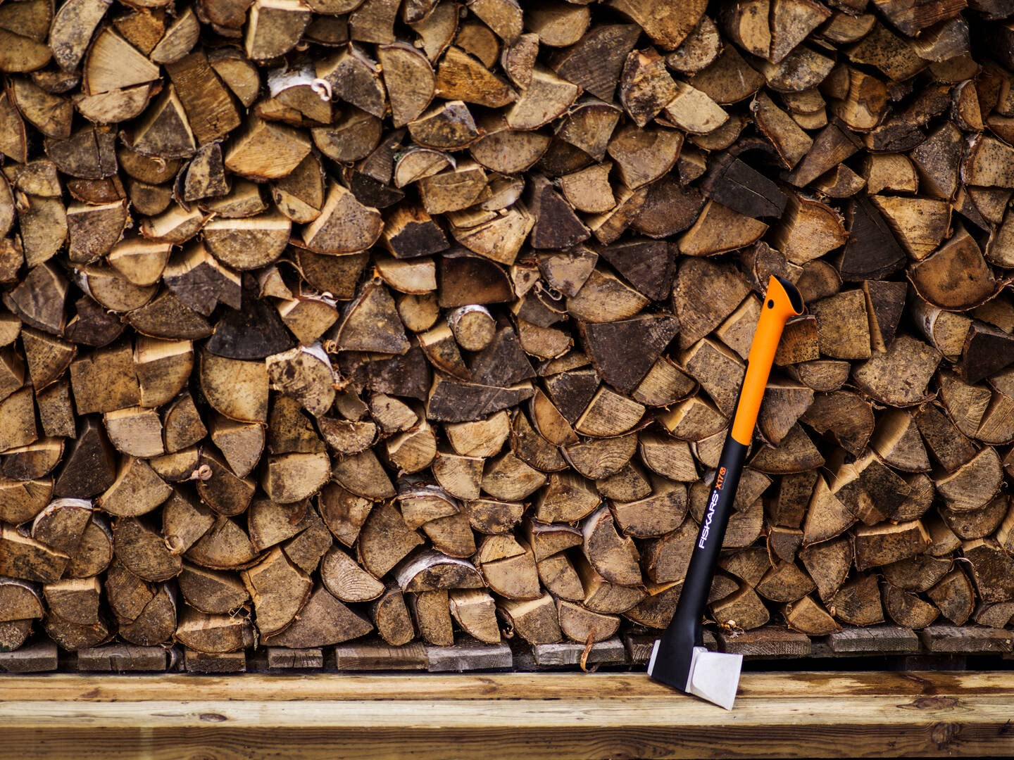 Eine orange-schwarze Axt lehnt vor einer großen Wand aus aufgestapelten Holzscheiten.