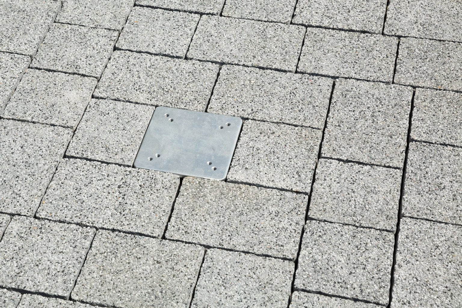 Eine quadratische Metallplatte ist in grauem Pflastersteinboden eingebettet.