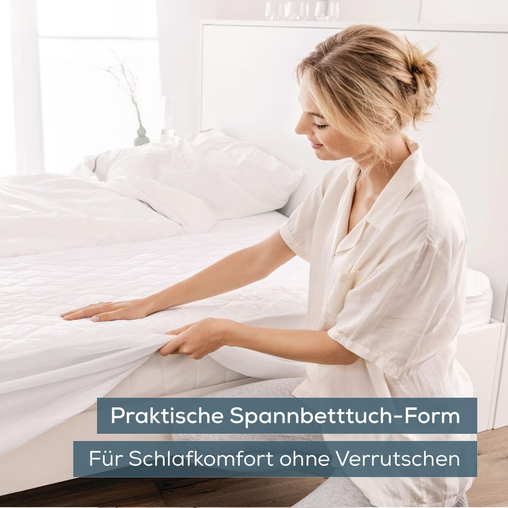 Una donna in abiti bianchi rifa il letto in una camera luminosa.