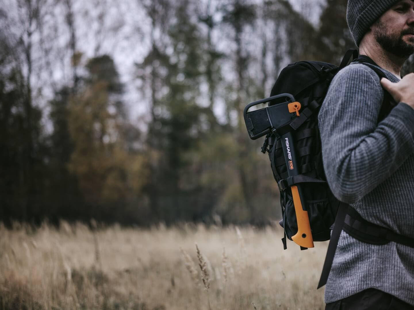 Un homme avec un sac à dos et une hache orange, debout dans un champ avec des arbres.