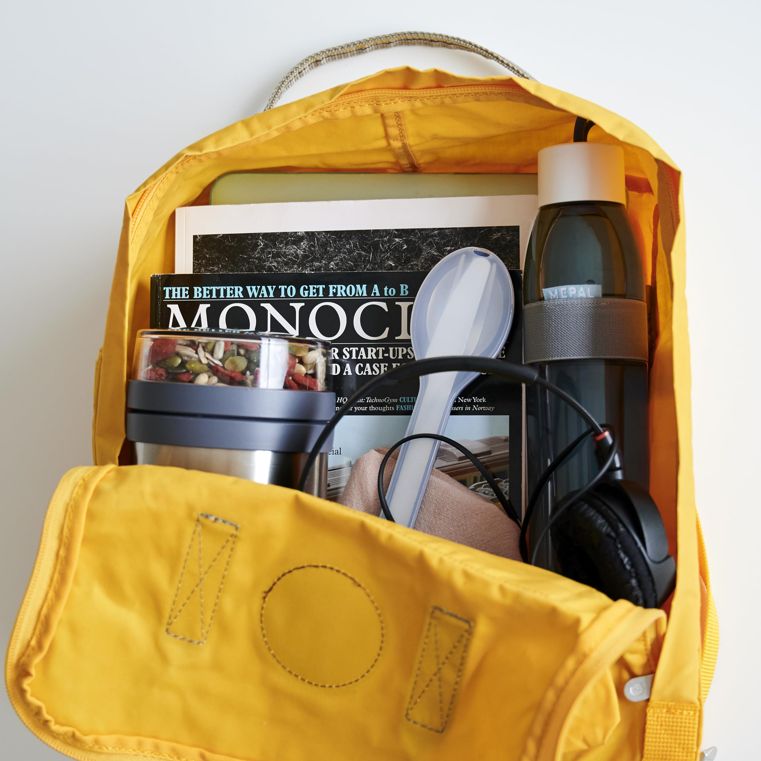 Zaino giallo aperto con rivista Monocle, portapranzo, borraccia, cuffie e cucchiaio.