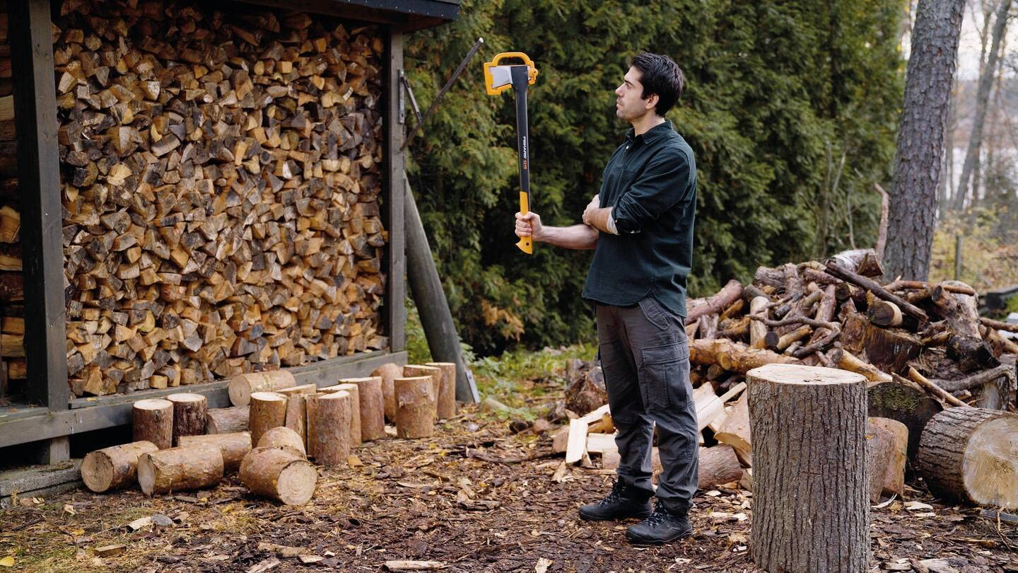 Ein Mann hält eine Axt vor einem Holzlager und einem Baumstumpf im Freien.