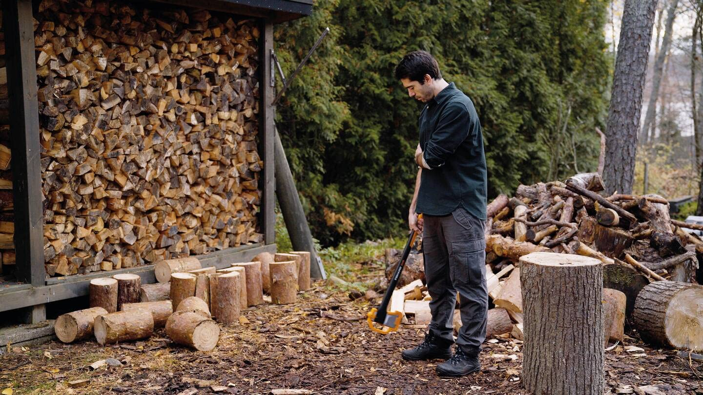 Ein Mann mit schwarzem Hemd steht mit einer Axt im Wald neben einem Holzstapel.