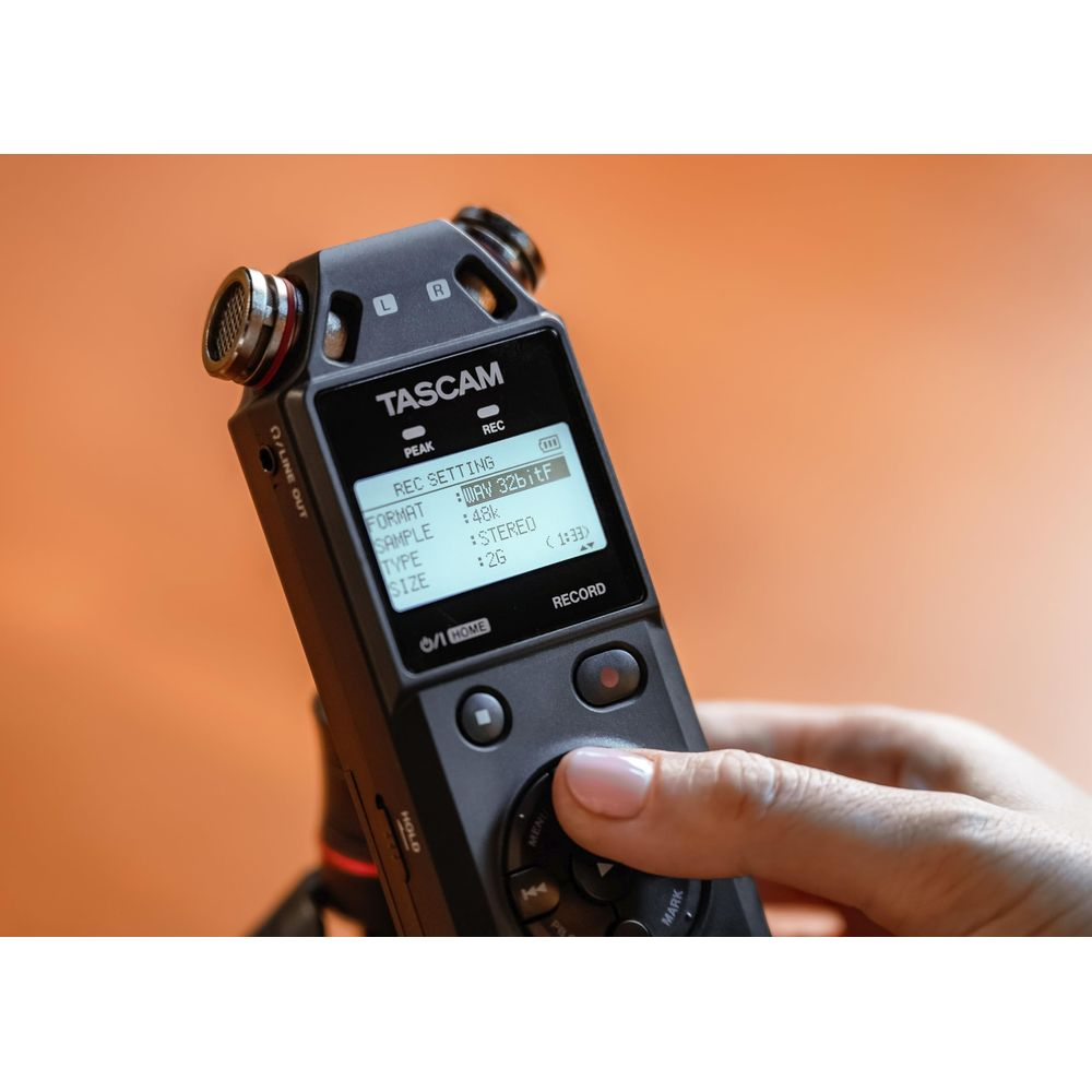 Une main manipule un enregistreur audio Tascam noir avec écran numérique sur fond orange.