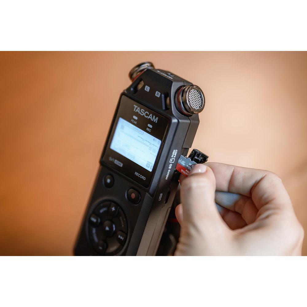 Une main insère une carte micro SD dans un enregistreur audio Tascam noir sur fond orange.