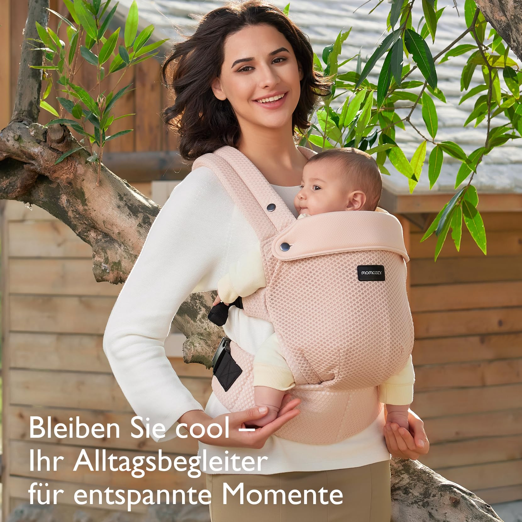 Una donna sorridente porta un bambino in un marsupio rosa davanti a una parete di legno.