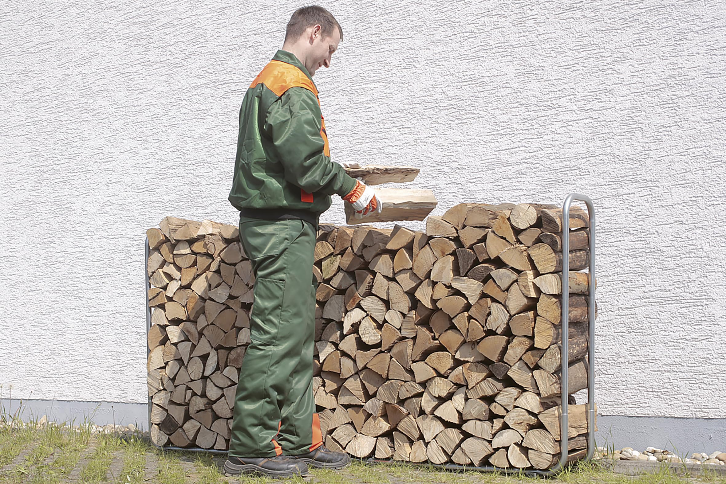 Un ouvrier en vêtements de travail verts empile du bois de chauffage dans un support en métal contre un mur blanc.