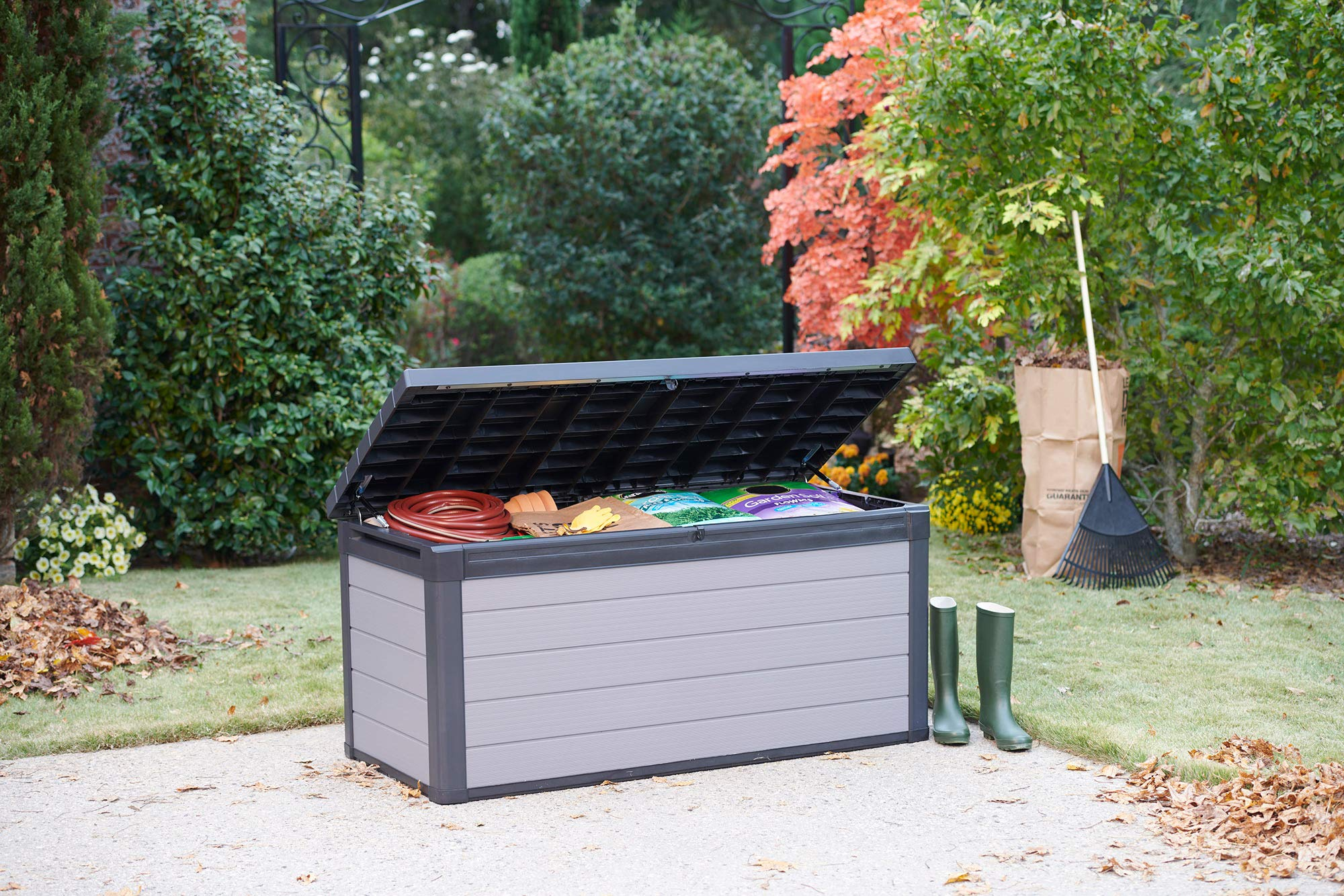 Eine offene grau-schwarze Gartenbox auf einer Terrasse, mit Gartengeräten in einem üppigen Garten.