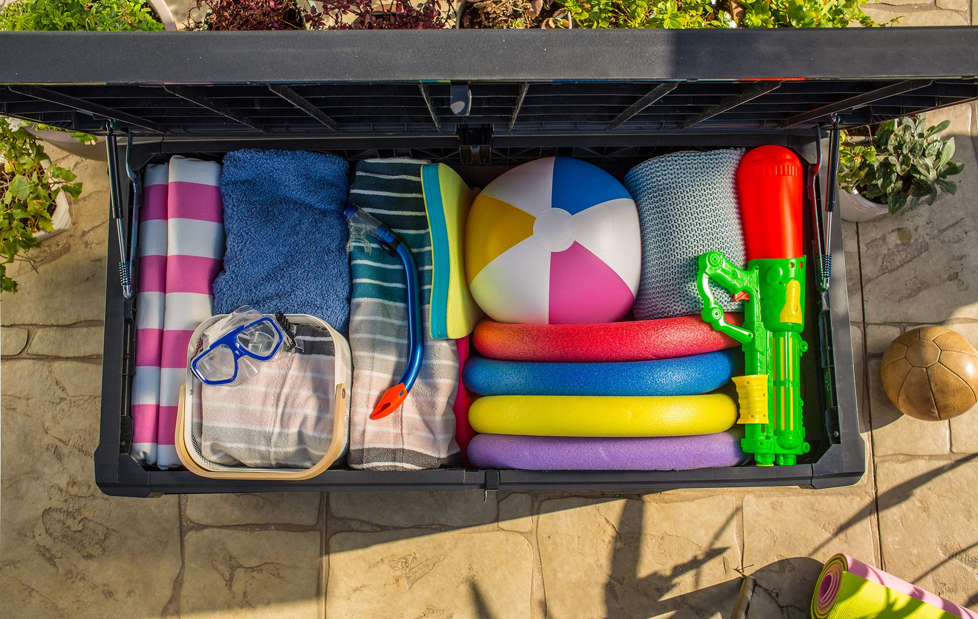 Eine geöffnete schwarze Kiste ist vollgepackt mit bunten Handtüchern, einem Strandball, Schwimmnudeln und Wasserspielzeug.