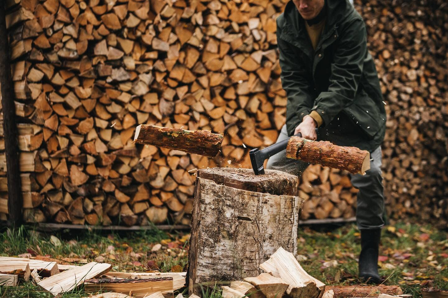 Un homme en veste verte fend une bûche avec une hache devant un grand tas de bois.
