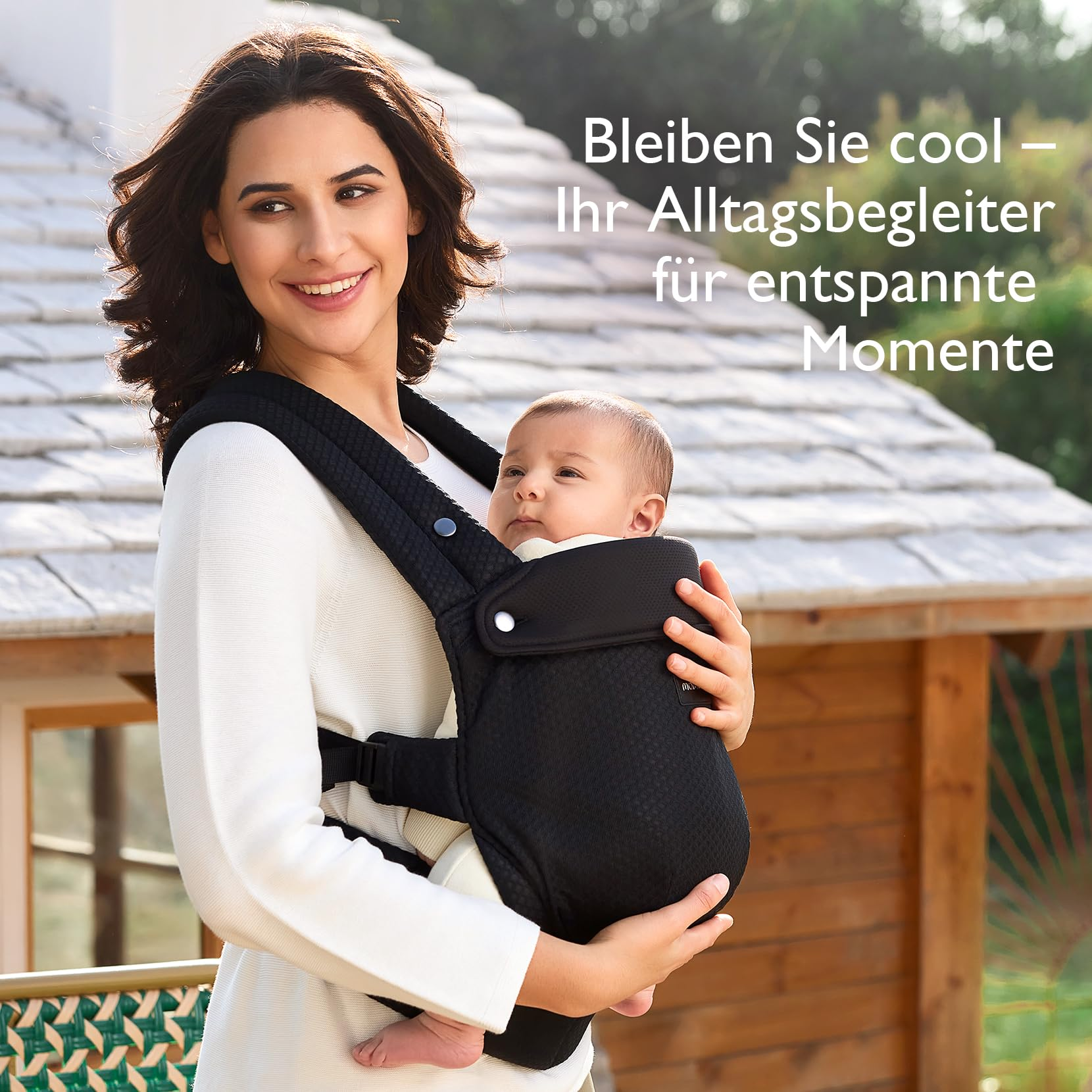 Una donna con un maglione bianco tiene un bambino in un marsupio nero davanti a una casa di legno.