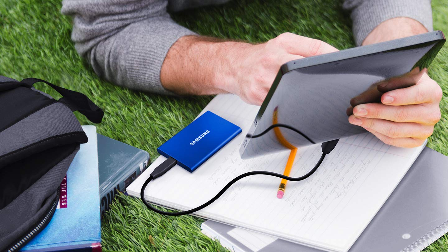 Una persona usa un tablet sull'erba, con un SSD esterno blu e uno zaino.