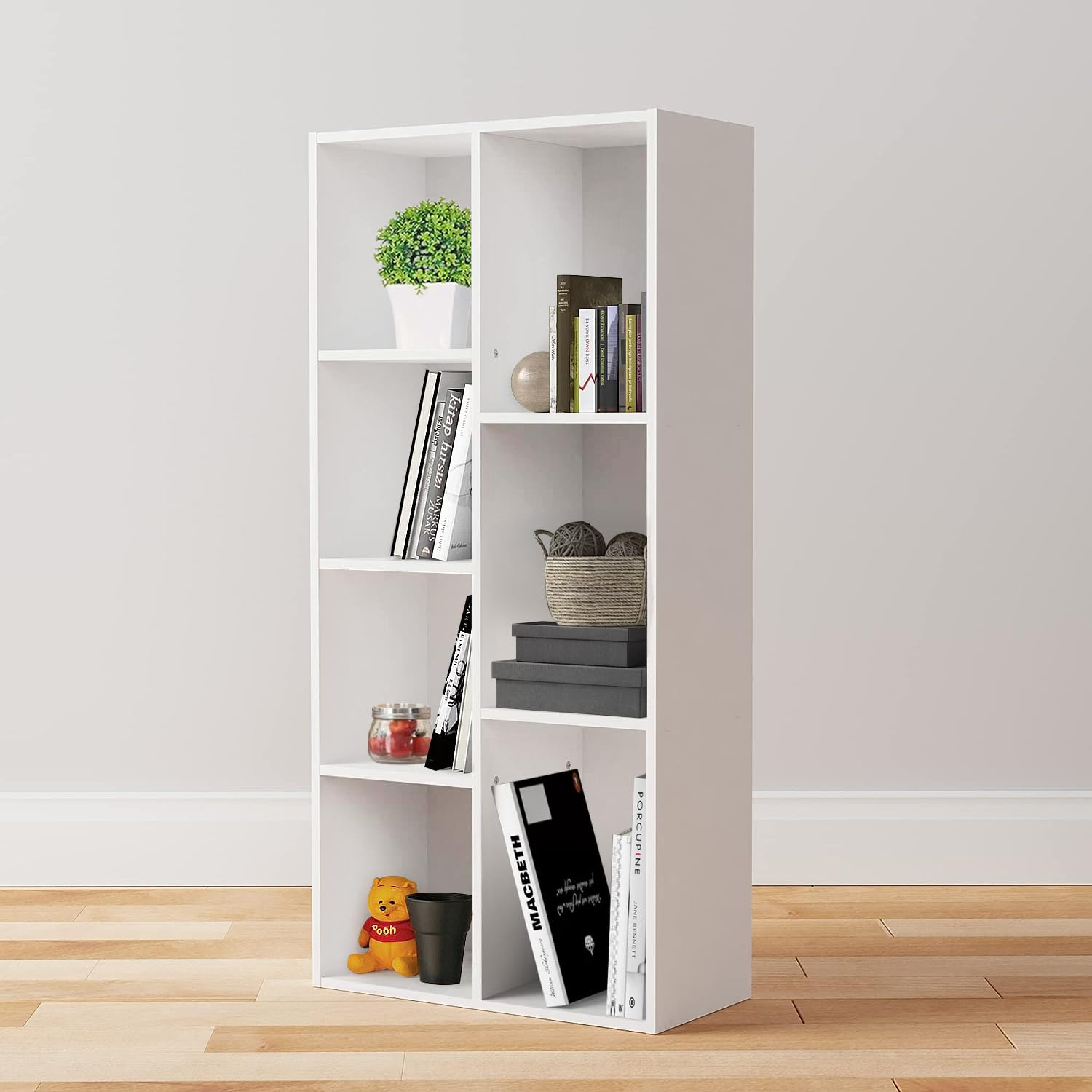 Une bibliothèque blanche à six compartiments sur parquet, décorée de livres, d'une plante et de petits objets.