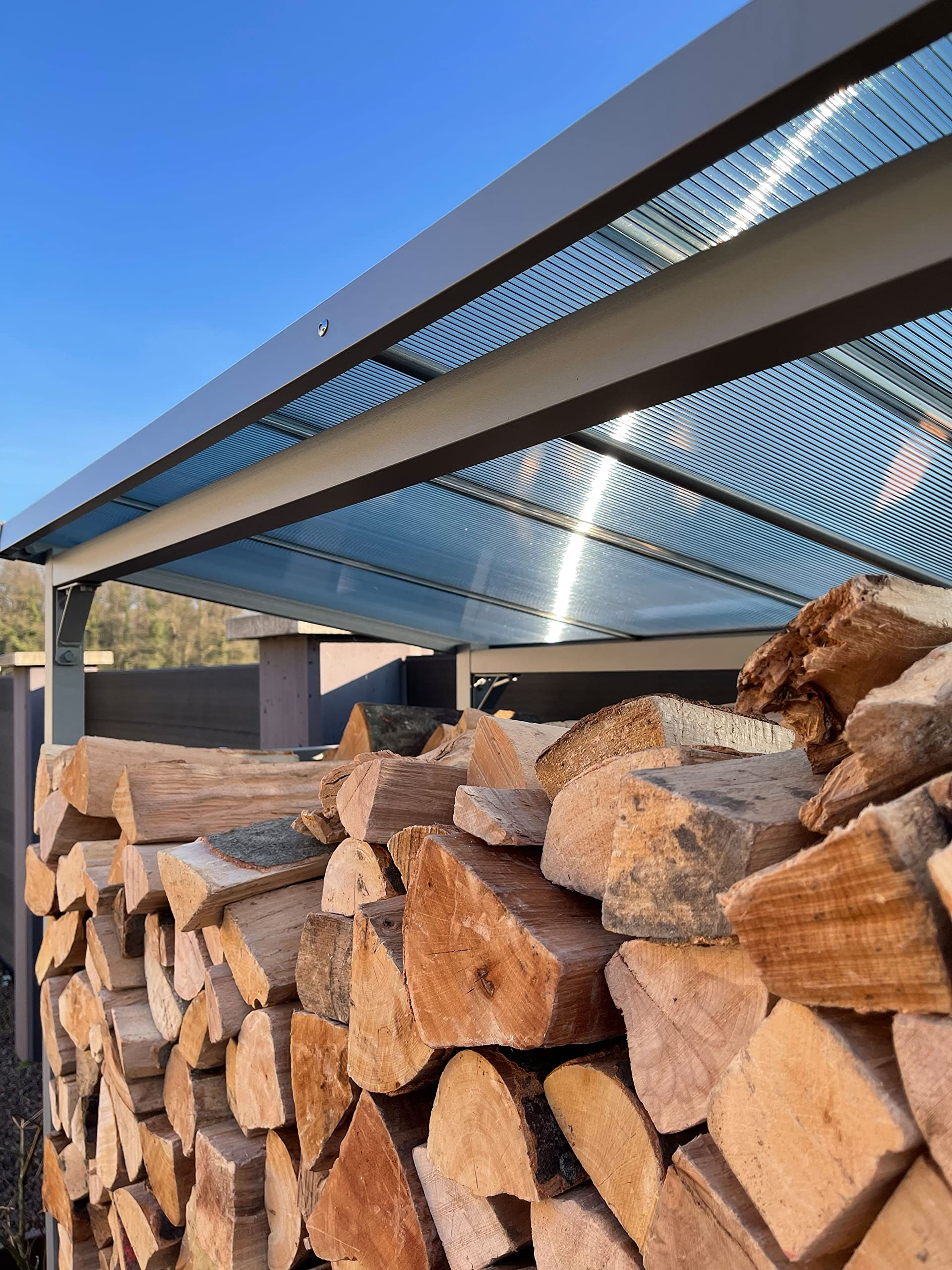 Pile de bois de chauffage sous un toit de terrasse en verre, ciel bleu.