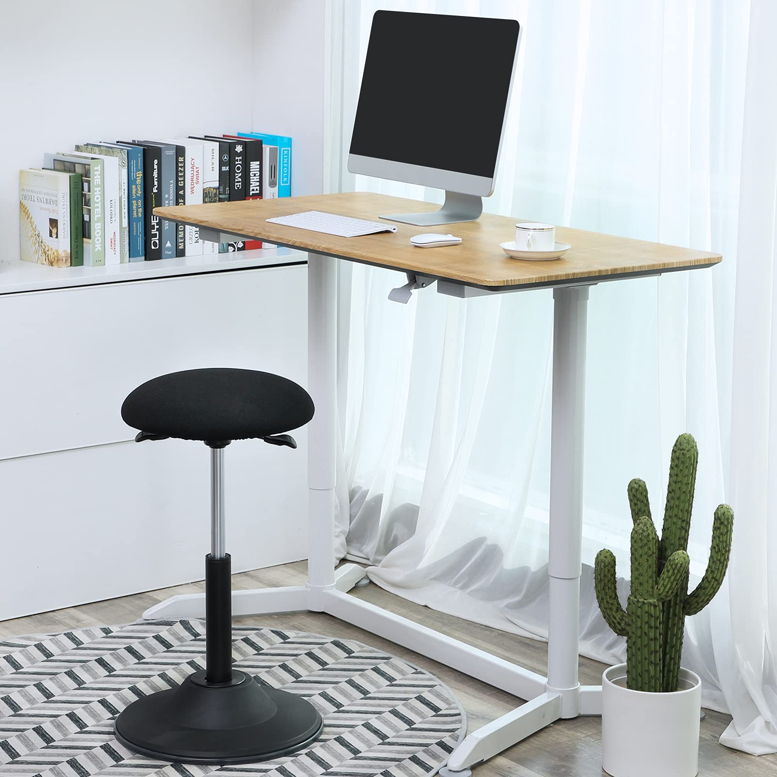 Un bureau réglable en hauteur avec ordinateur, tabouret, livres et un cactus sur un tapis à motifs.
