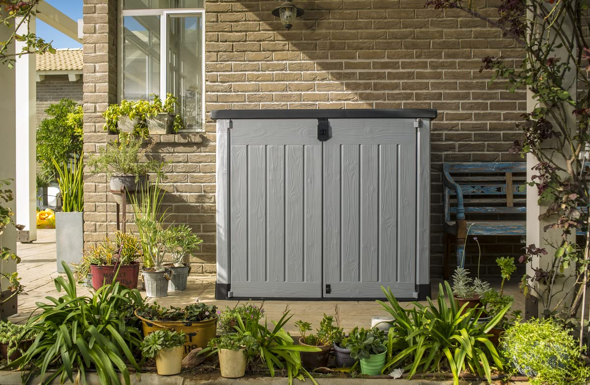 Grauer Kunststoff-Gartenschrank vor einer Ziegelwand, umgeben von Topfpflanzen auf einer Terrasse.