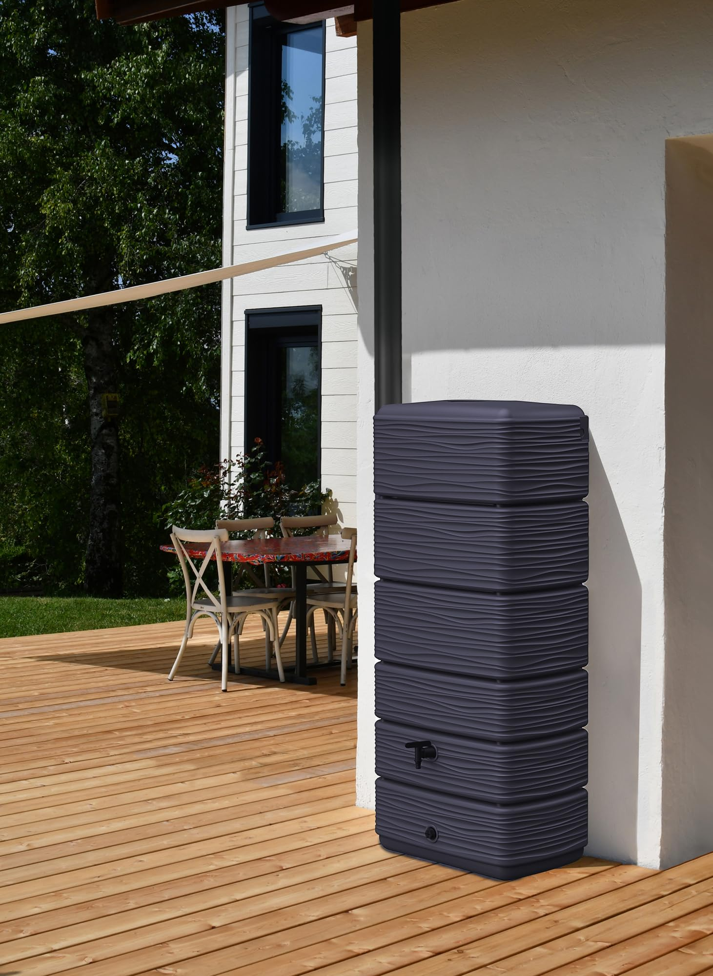 Récupérateur d'eau de pluie gris foncé sur une terrasse en bois devant une maison blanche.