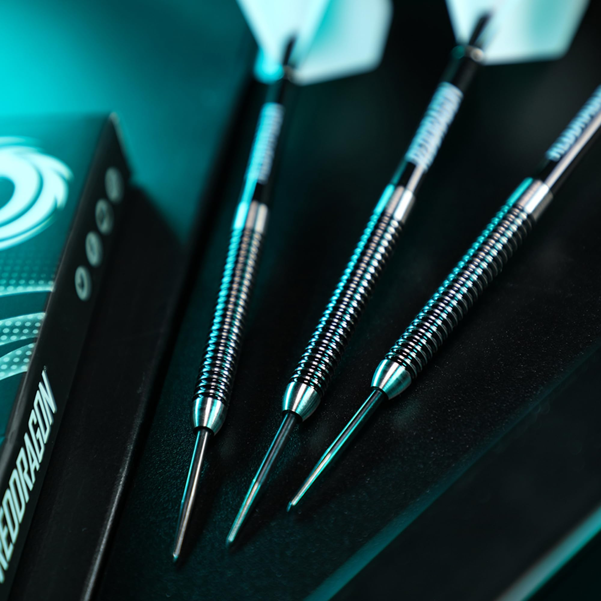 Drei silberne Darts mit gerillten Barrels und weißen Flights liegen nebeneinander neben einer schwarzen Box in tealem Licht.