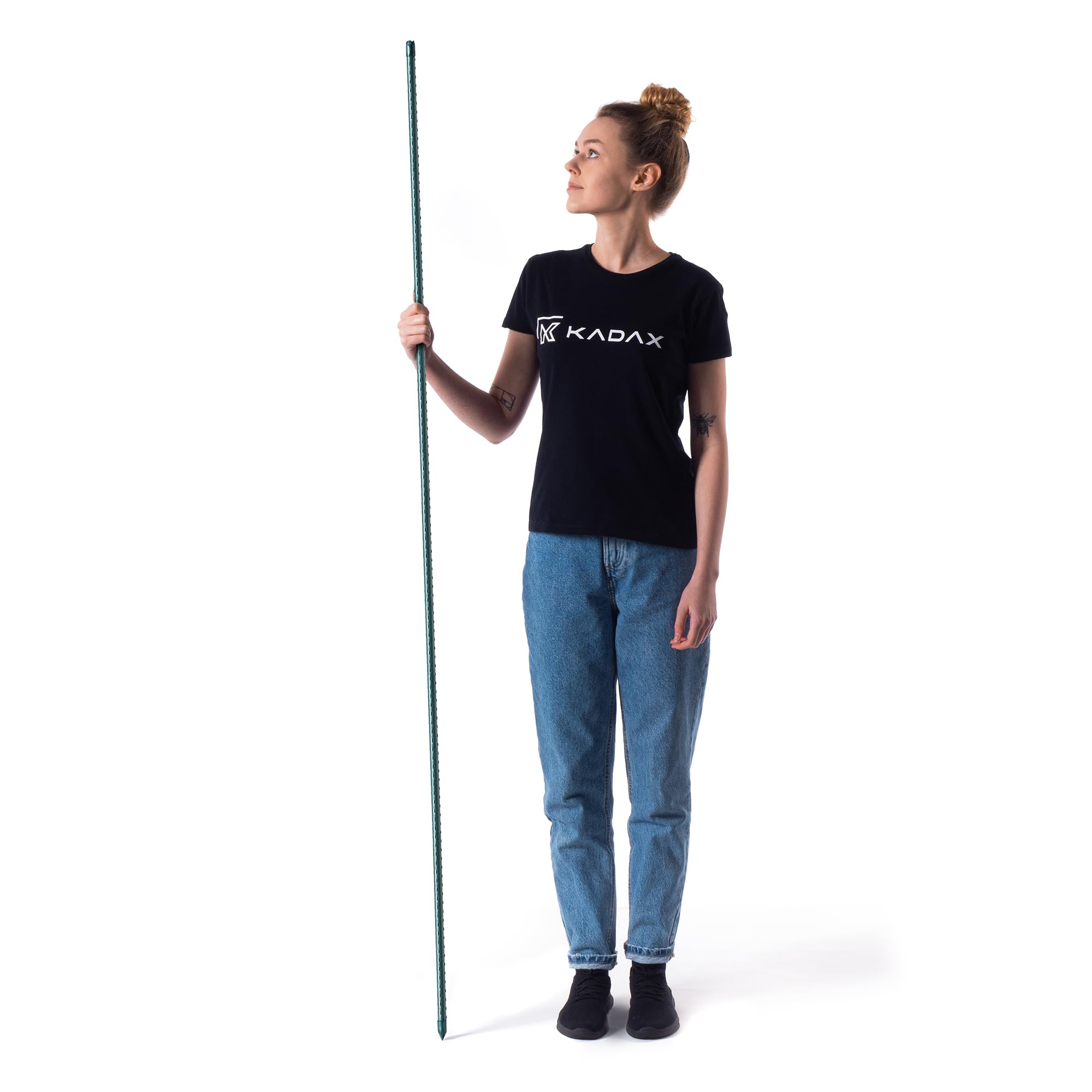 Une femme blonde en t-shirt KADAX noir et jean bleu tient un long bâton vert sur fond blanc.