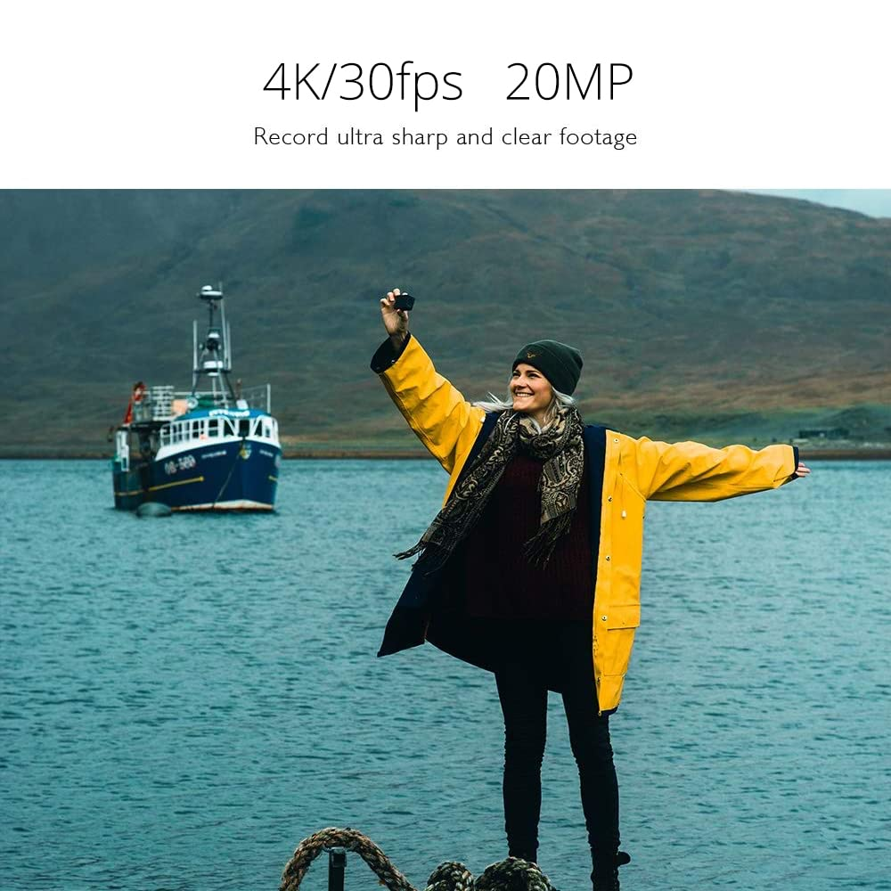 Une femme en veste jaune tient un appareil photo près de l'eau avec un bateau en arrière-plan.