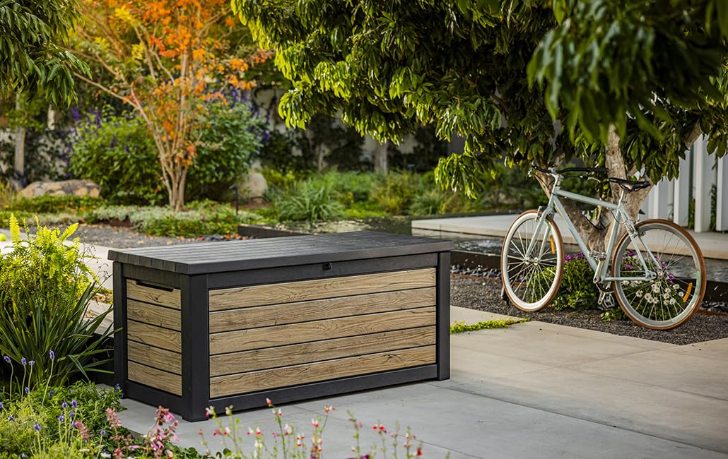 Eine dunkelgraue und holzverkleidete Aufbewahrungsbox auf einer Terrasse im Garten, daneben ein Fahrrad.