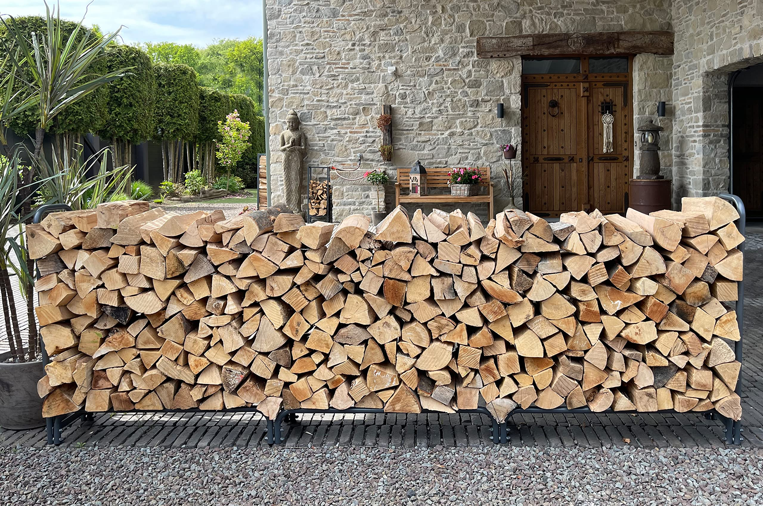 Une pile de bois de chauffage fendu dans un support en métal devant un mur en pierre, avec porte et jardin.