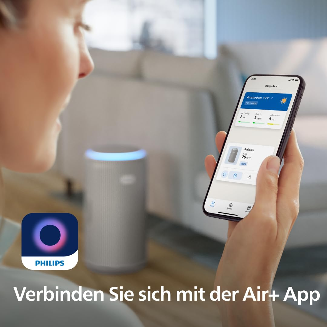 Eine Person hält ein Smartphone mit der Philips Air+ App, im Hintergrund ein Luftreiniger im unscharfen Wohnzimmer.