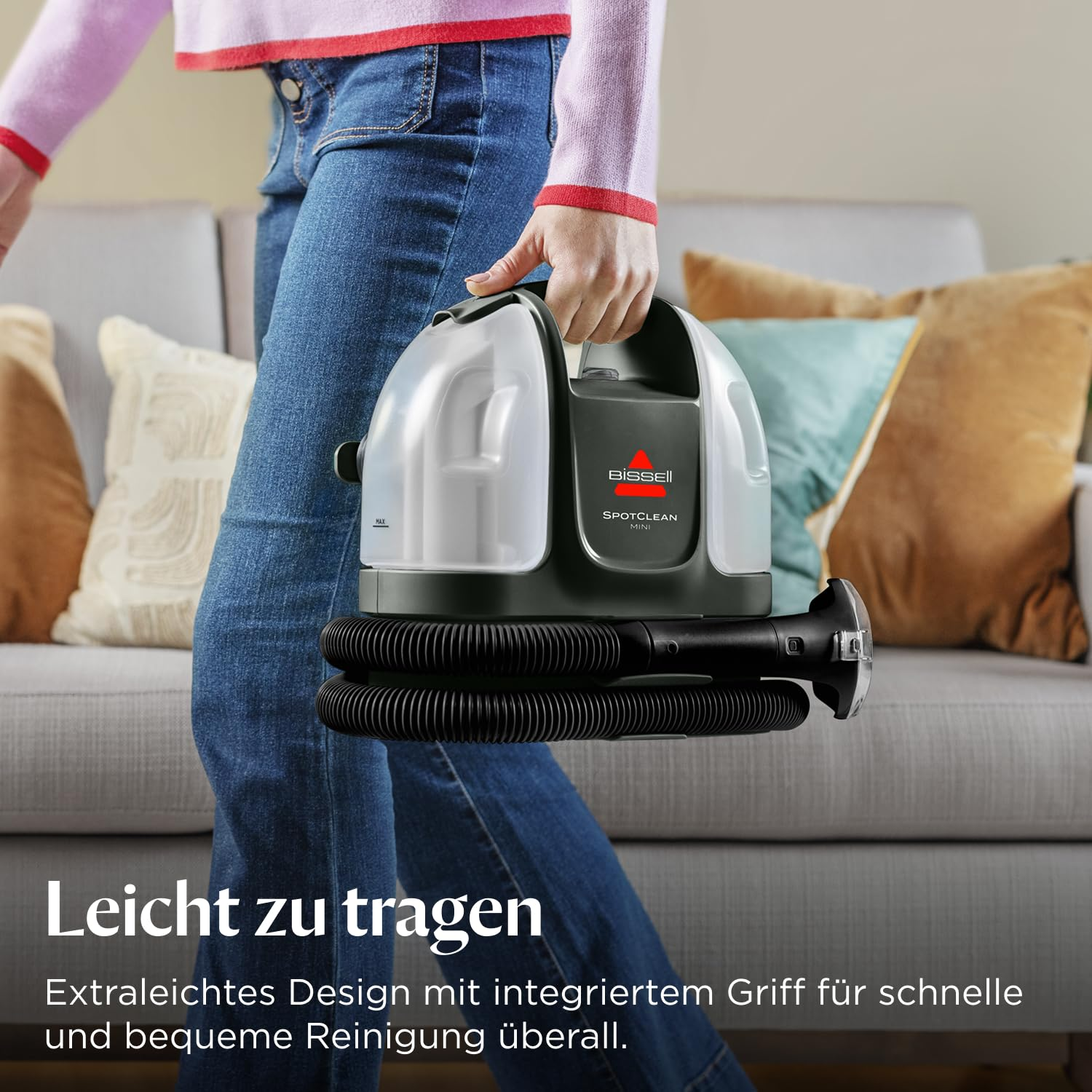 Persona in jeans trasporta un pulitore grigio Bissell SpotClean Mini davanti a un divano con cuscini.