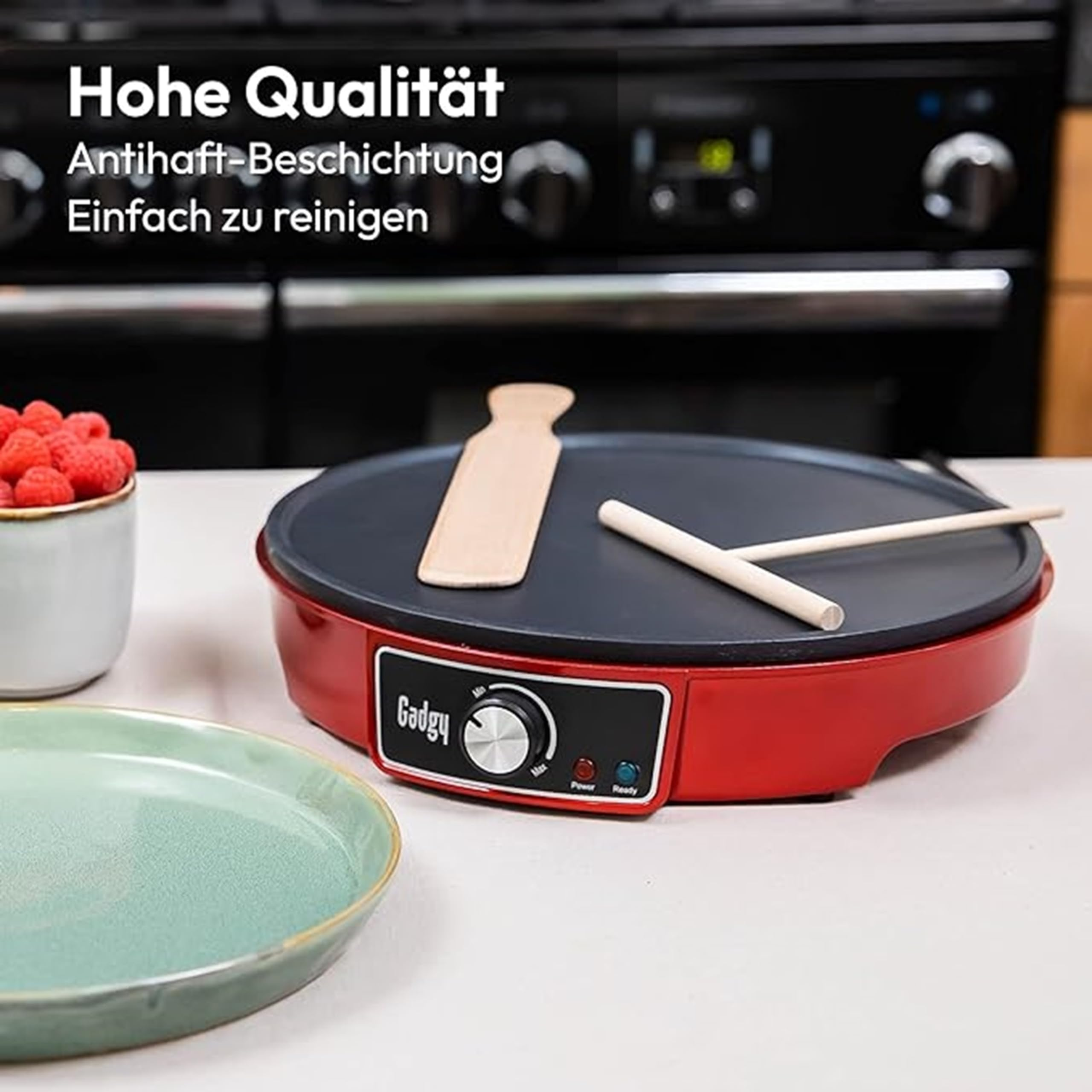 Crépière électrique rouge avec ustensiles en bois, une assiette et des framboises sur un comptoir.