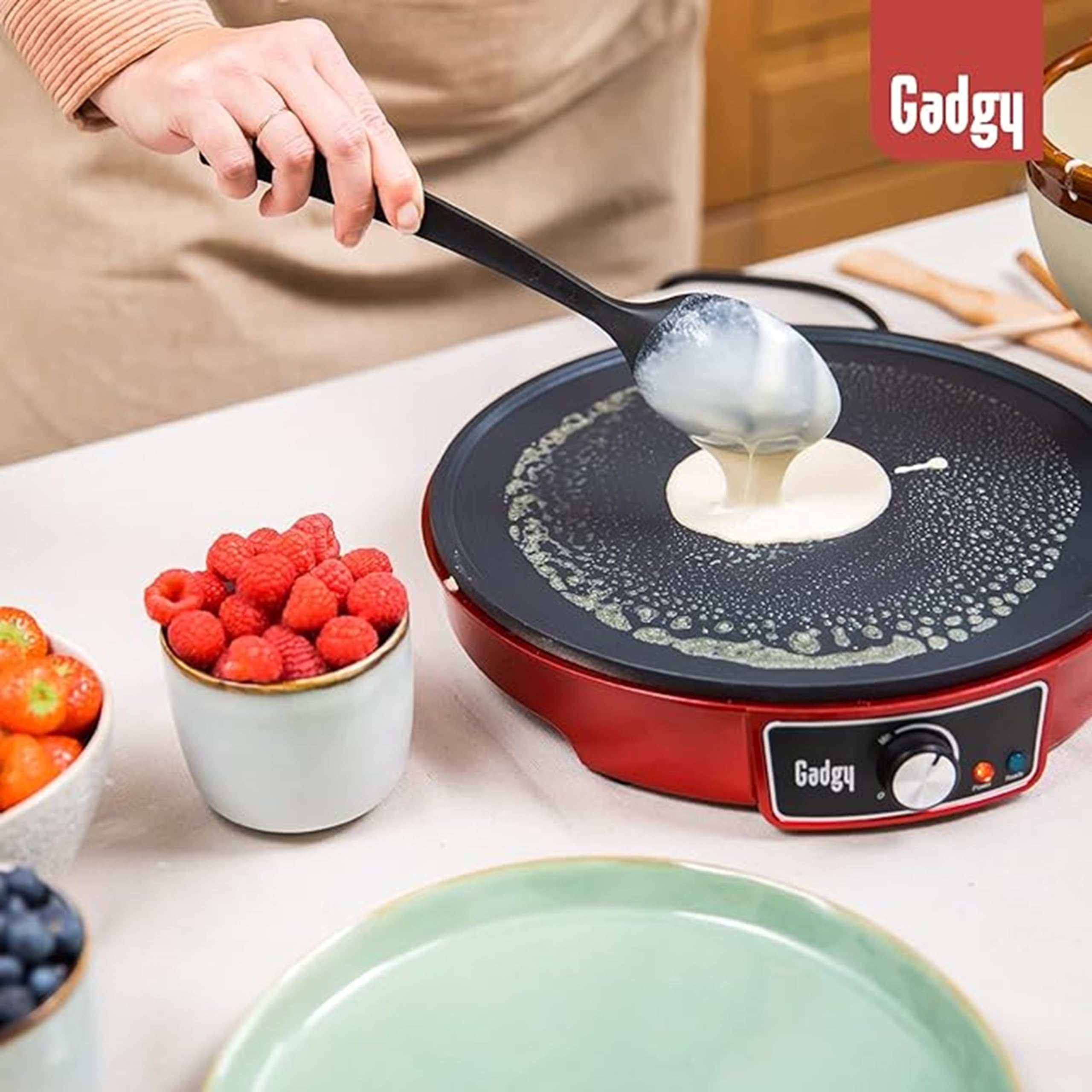 Une personne verse de la pâte avec une louche sur une crêpière ronde rouge. Des framboises fraîches sont à côté.