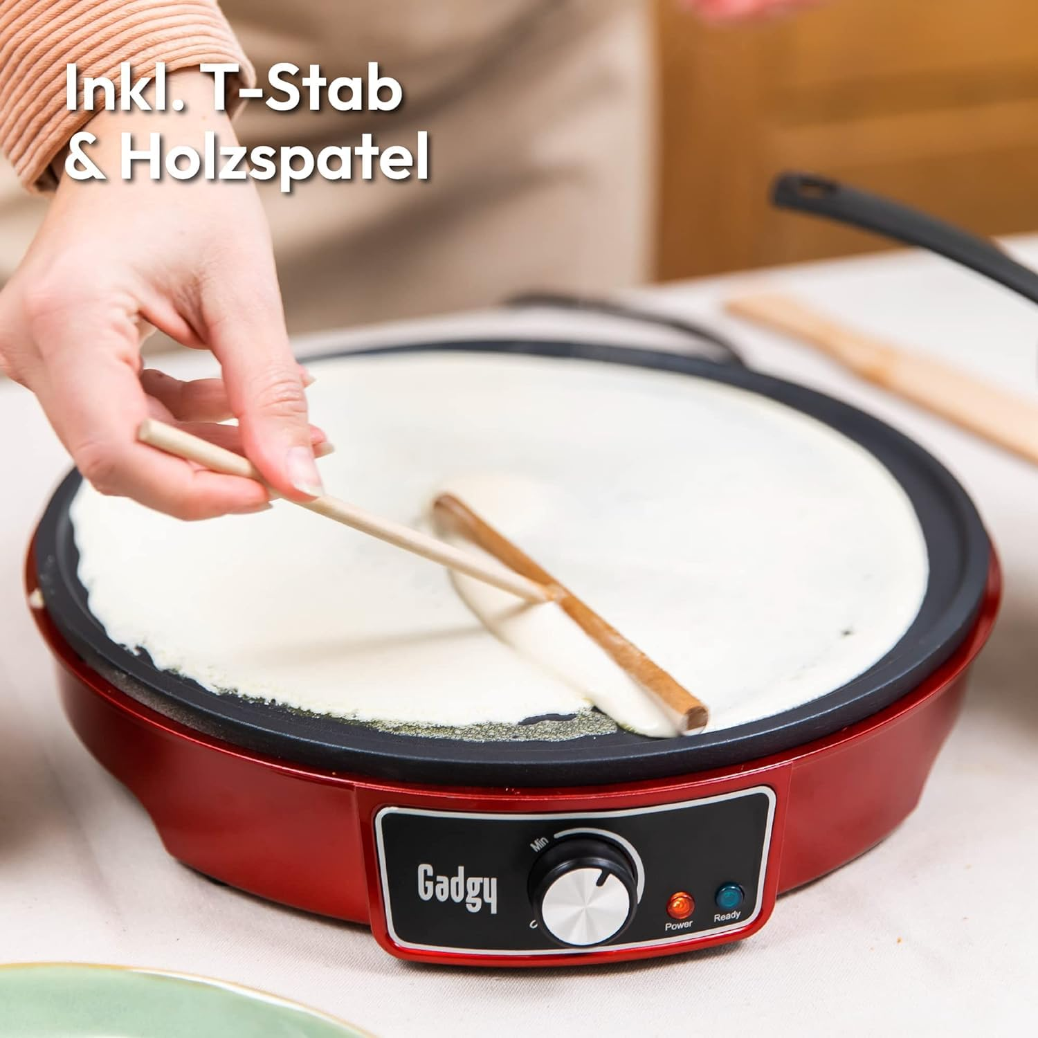 Une main utilise un bâton en T en bois pour étaler la pâte sur une crêpière électrique rouge.