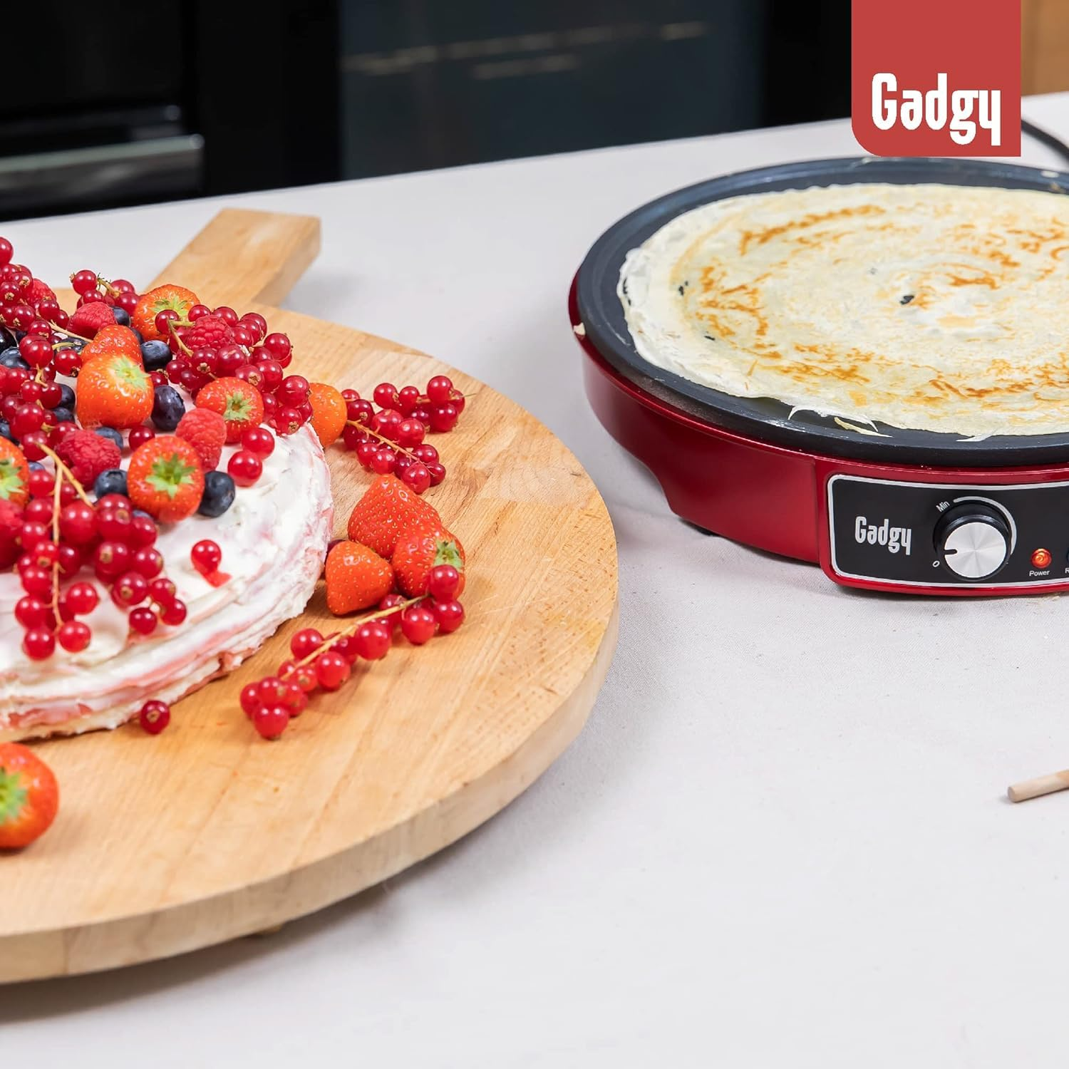 Une crêpière cuit une crêpe à côté d'une planche en bois avec un gâteau et des baies fraîches.