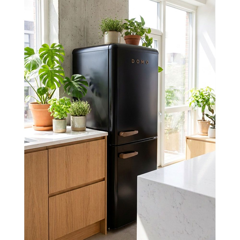 Un réfrigérateur rétro noir se trouve dans une cuisine lumineuse, entouré de plusieurs plantes vertes et de meubles en bois.