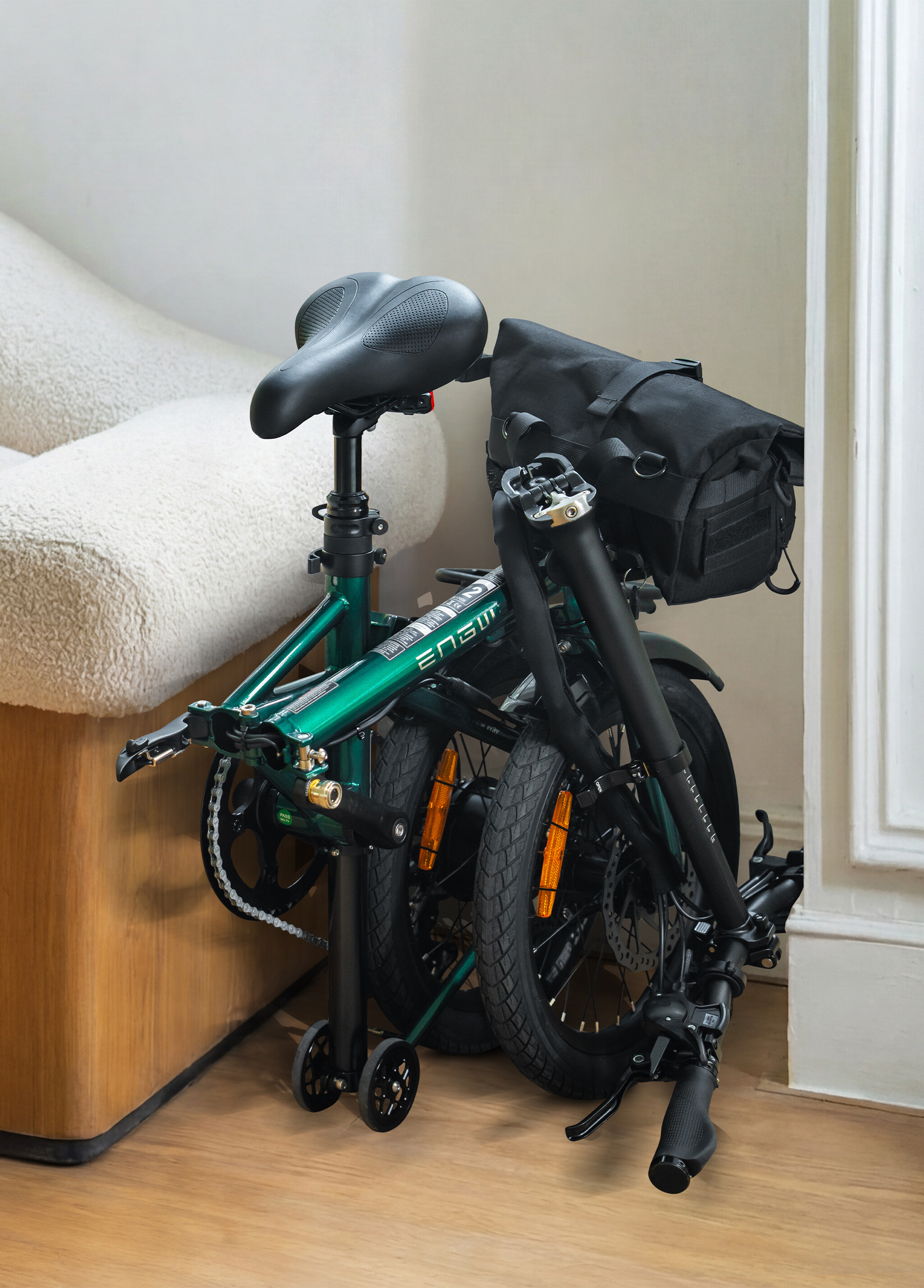 Una bicicletta pieghevole verde e nera chiusa con una borsa nera si trova all'interno su un pavimento in legno.
